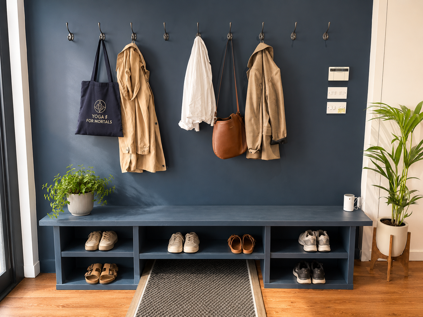 Entryway with a blue wall and a shoe storage bench, featuring sneakers, sandals, and black shoes underneath. Coats and bags hanging on hooks, left to right: navy tote bag, beige trench coat, white shirt, brown leather crossbody bag, and checked blaze