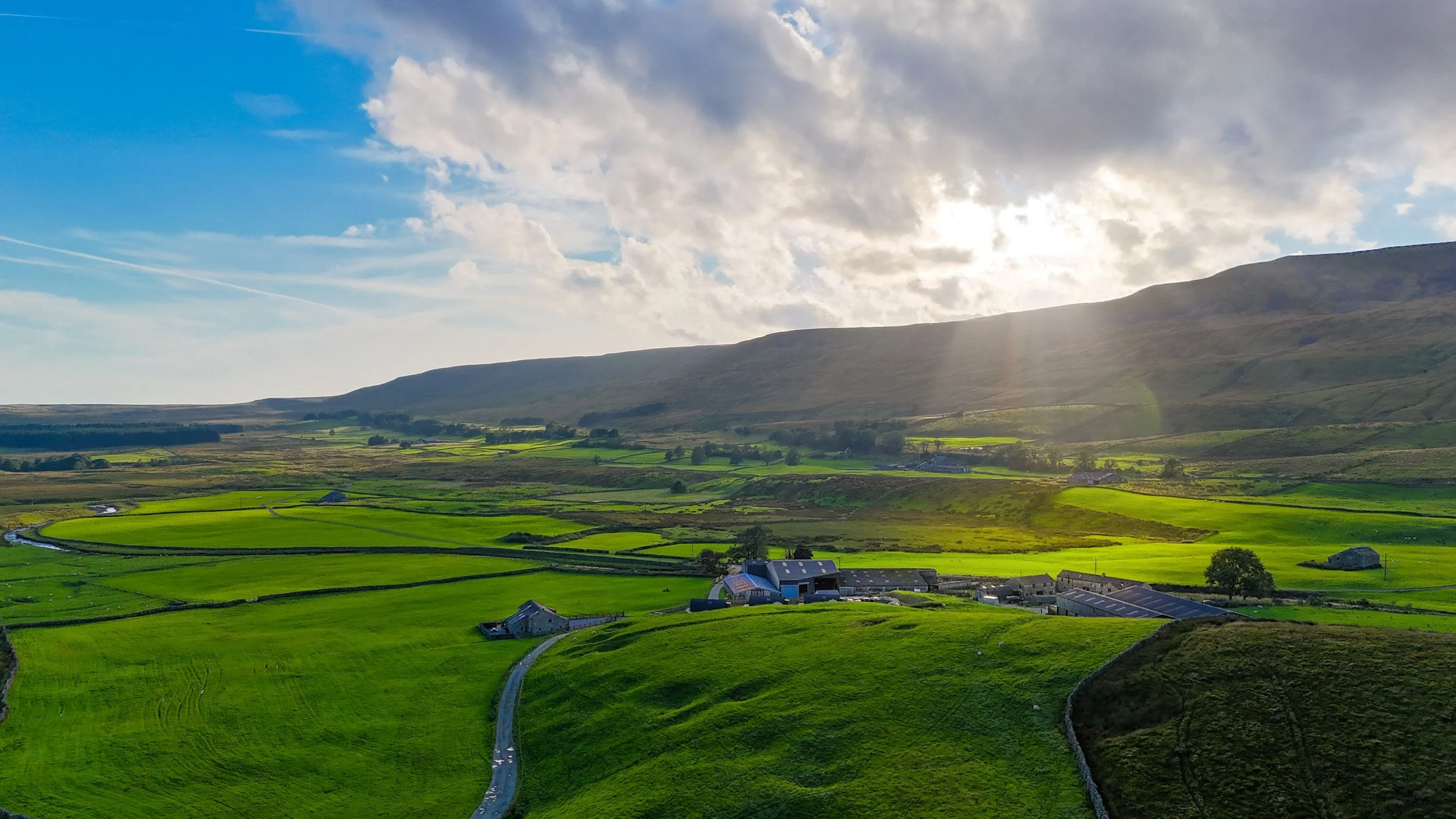 A scenic landscape of green fields, farm buildings, a winding road, rolling hills, and a mountain range in the distance under a partly cloudy sky with sunlight breaking through.