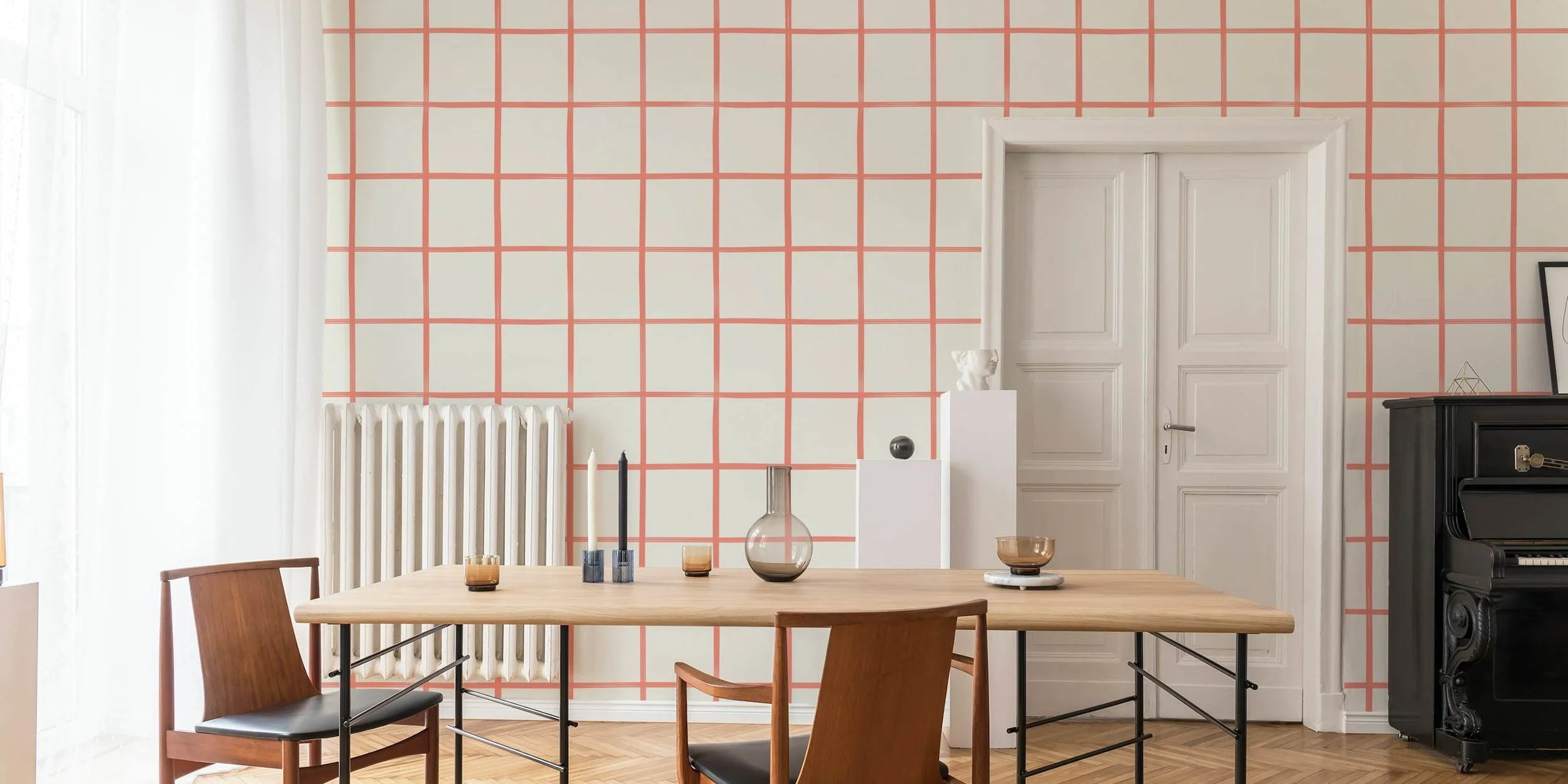 A dining room with a wooden table, modern chairs, a white radiator, and a patterned wall with red grid lines. Candles and vases are on the table, a white door is in the background, and a black piano is on the right side.