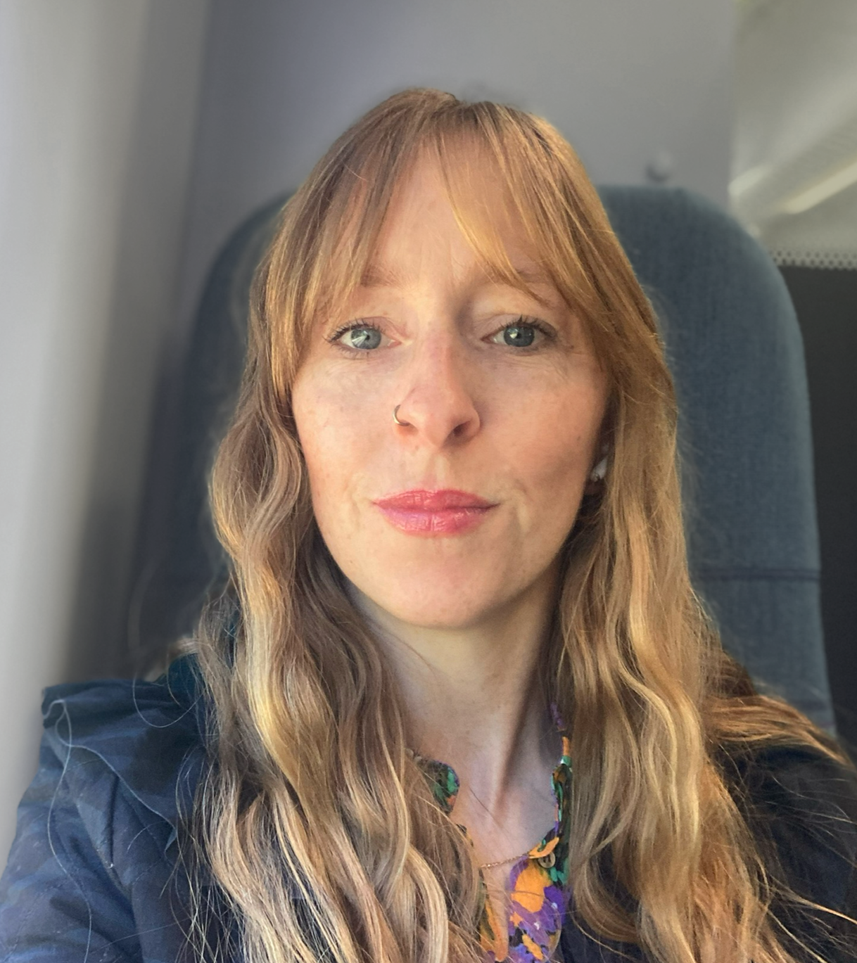 A woman with long, wavy, light brown hair and blue eyes sitting in what appears to be an airplane seat, wearing a navy blue top and a colorful necklace.