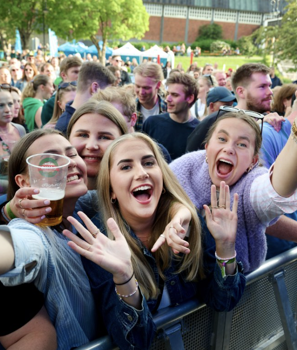 Grupp unge voksne som deler et øyeblikk av glede og moro på en utendørs festival, mange smil og lykke, med en barriere foran og et grønt område bak.