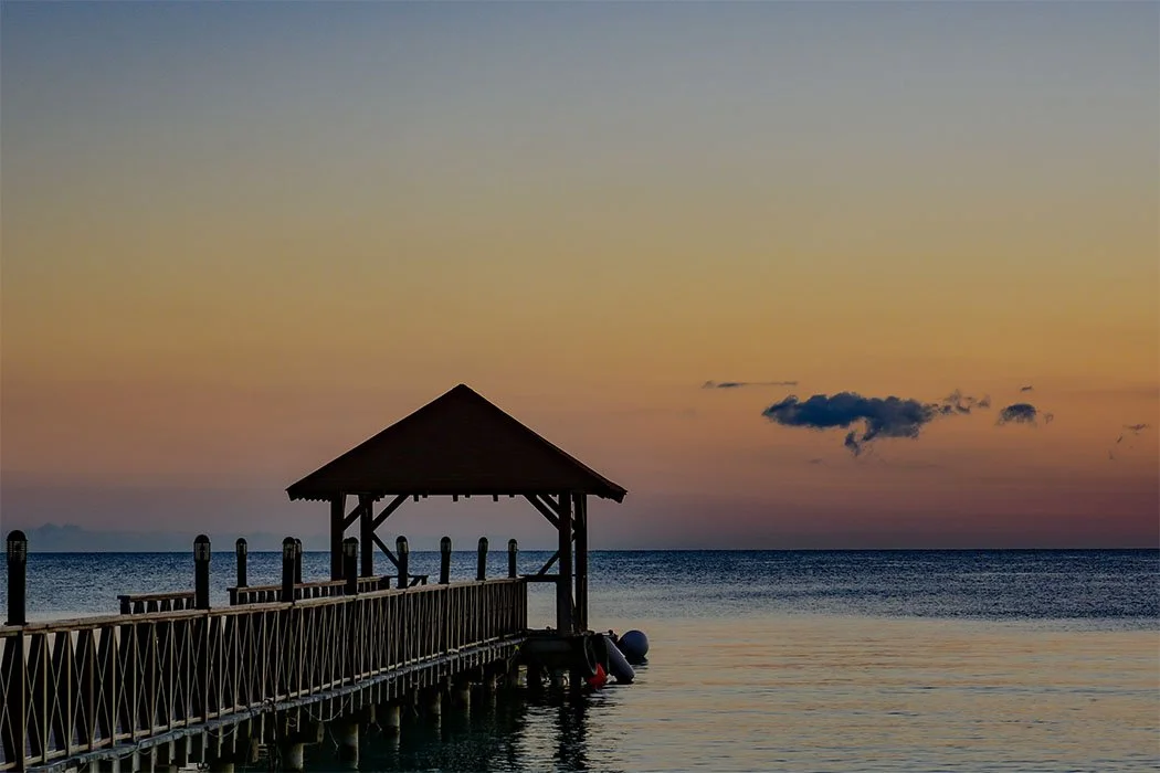 A wooden pier with a small gazebo extending over calm water at sunset, with a colorful sky and a few clouds.