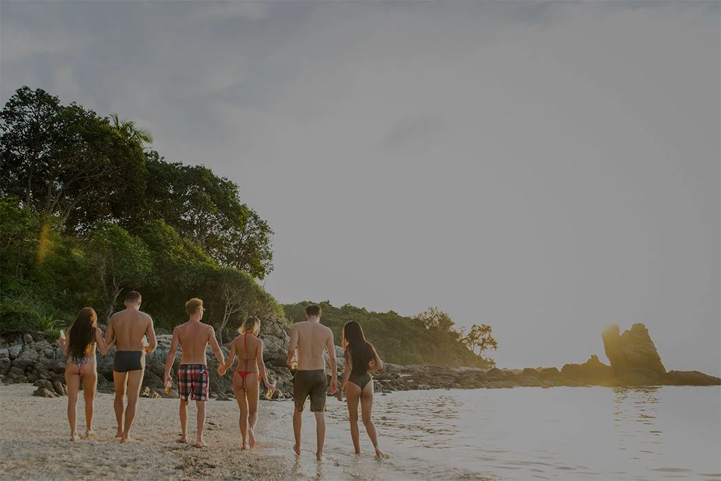 Six young people in swimsuits walking hand in hand along a beach at sunset, with rocks and trees in the background.