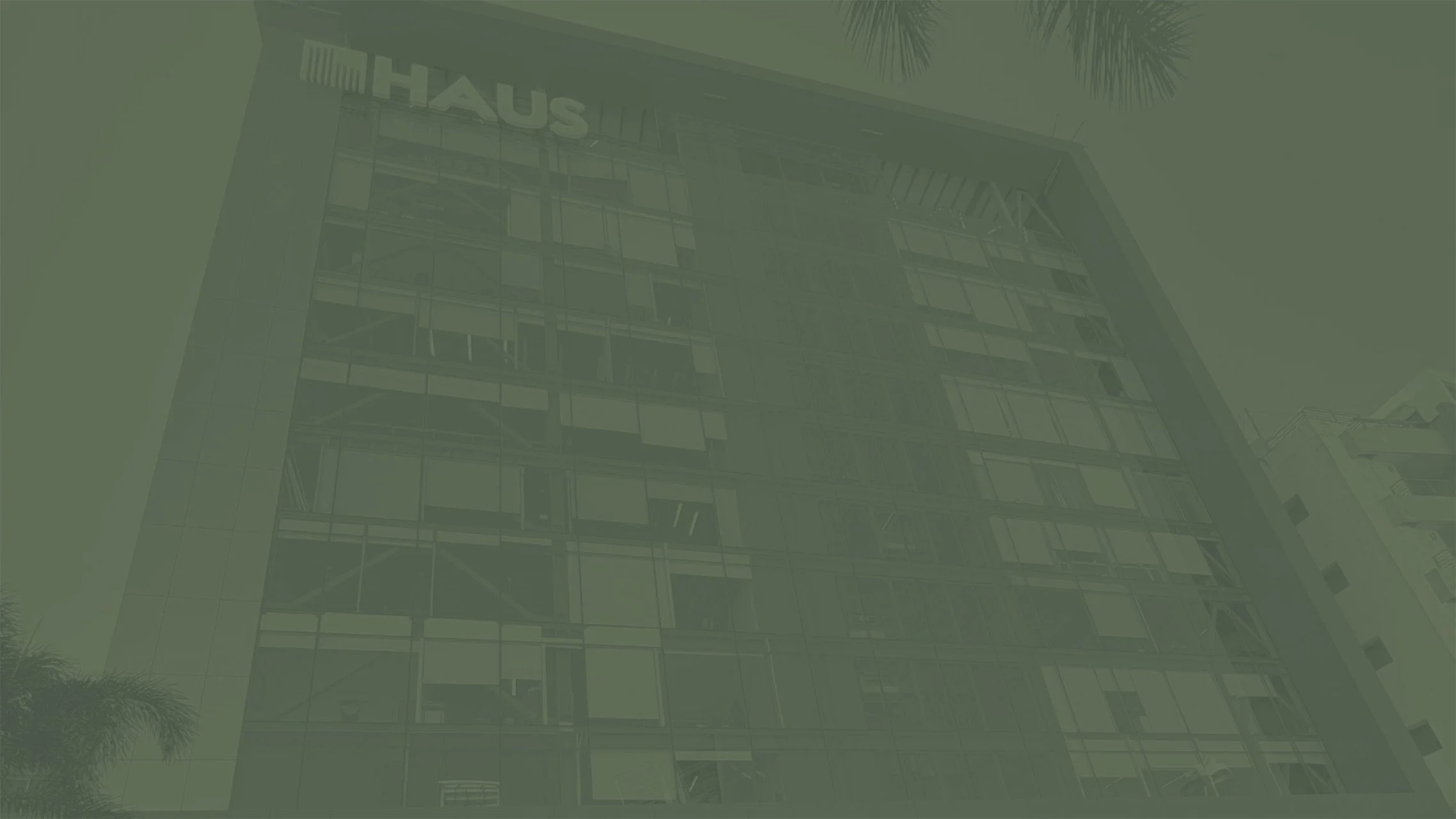 Glass-fronted building with the sign "Haus" at the top, palm trees nearby, and reflecting windows.