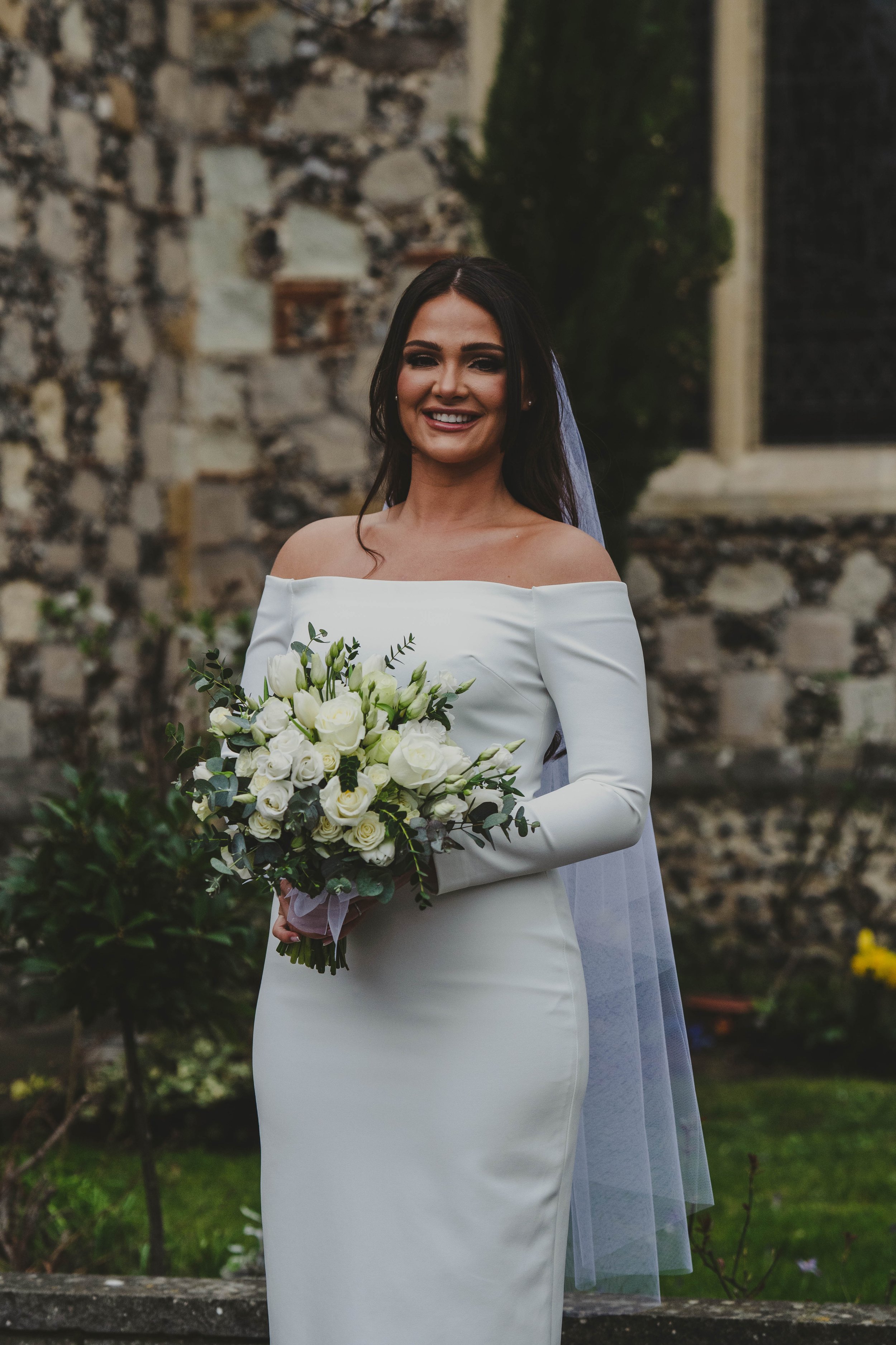 A smiling woman in a white off-shoulder wedding dress holding a bouquet of white roses, standing outdoors with a stone wall and greenery in the background.