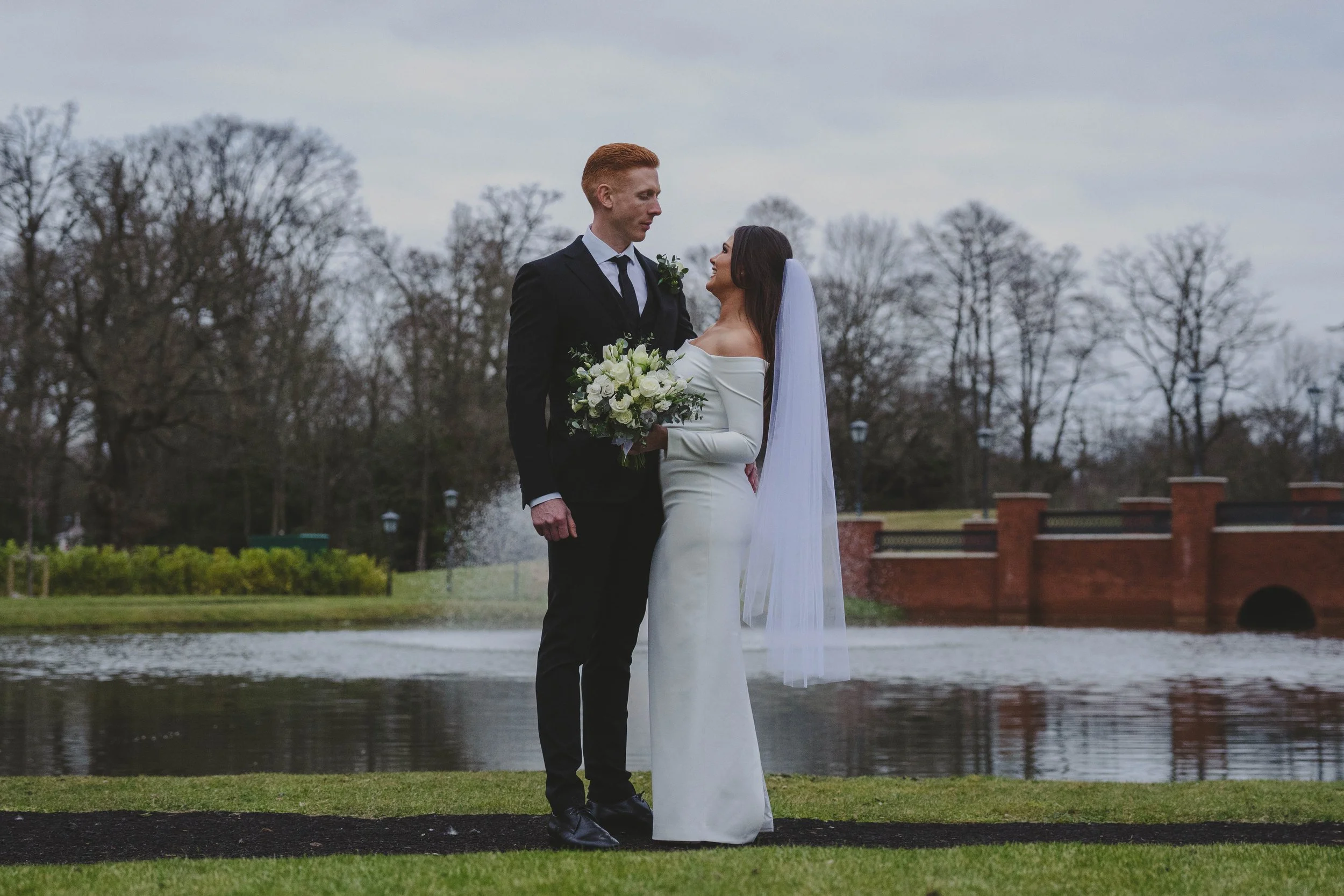 A bride and groom holding hands and gazing at each other outdoors near a lake with a fountain, trees, and a brick bridge in the background.