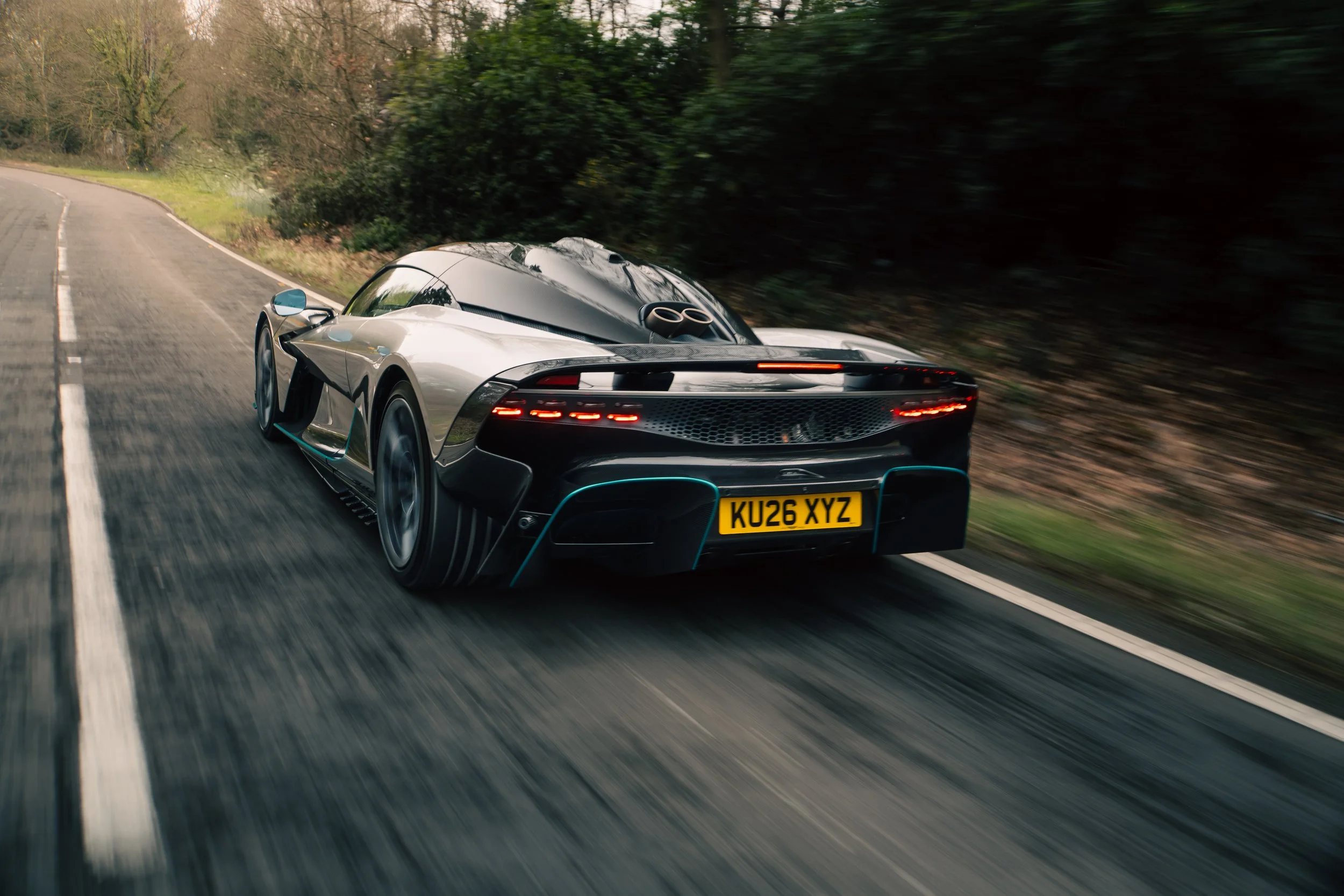 A sleek silver sports car with a black roof speeding along a winding rural road surrounded by trees.