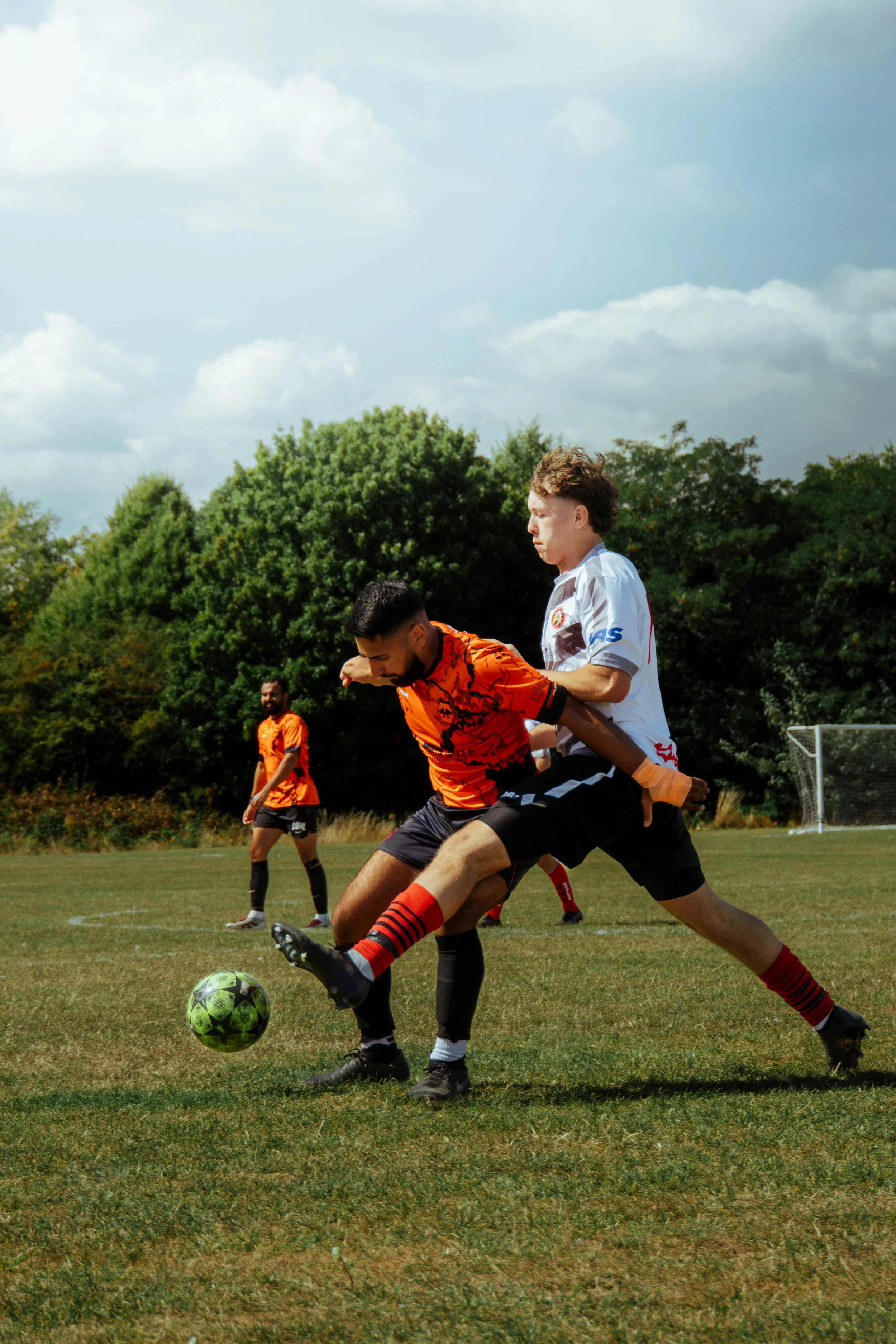 Two soccer players competing for the ball during a match on a grassy field with a few other players visible in the background, trees, and a cloudy sky.