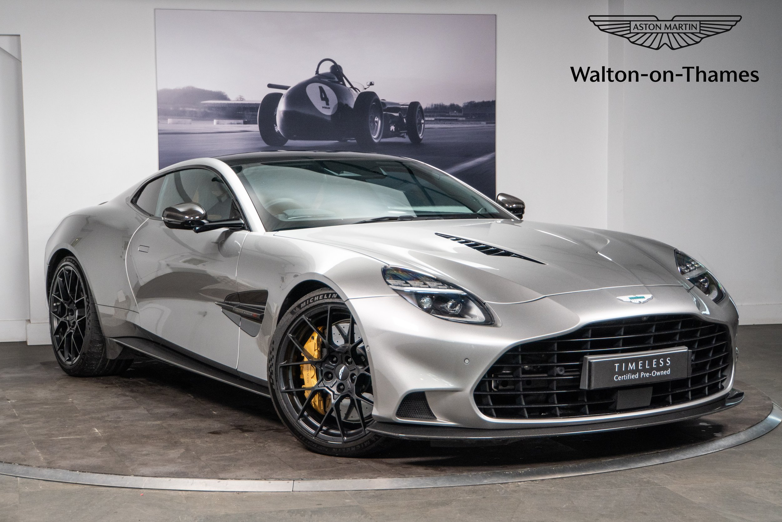 A silver Aston Martin sports car on display at a showroom, with a black and white racing car poster behind it and a black sign that reads "Timeless" and "Certified Pre-Owned."