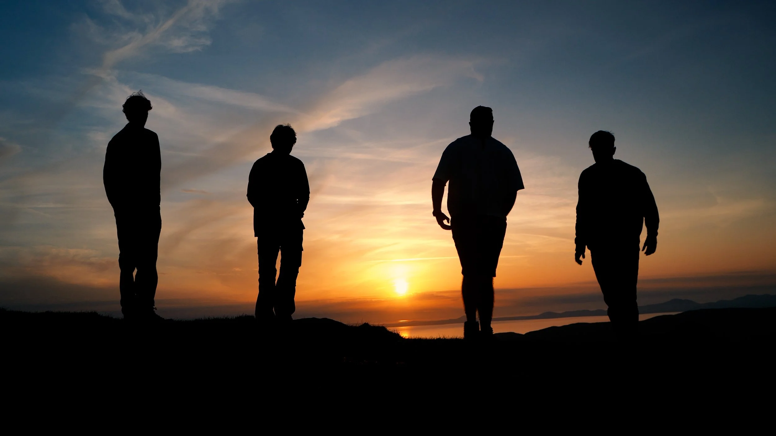 Silhouettes of four people walking outdoors at sunset with a colorful sky and water in the background.