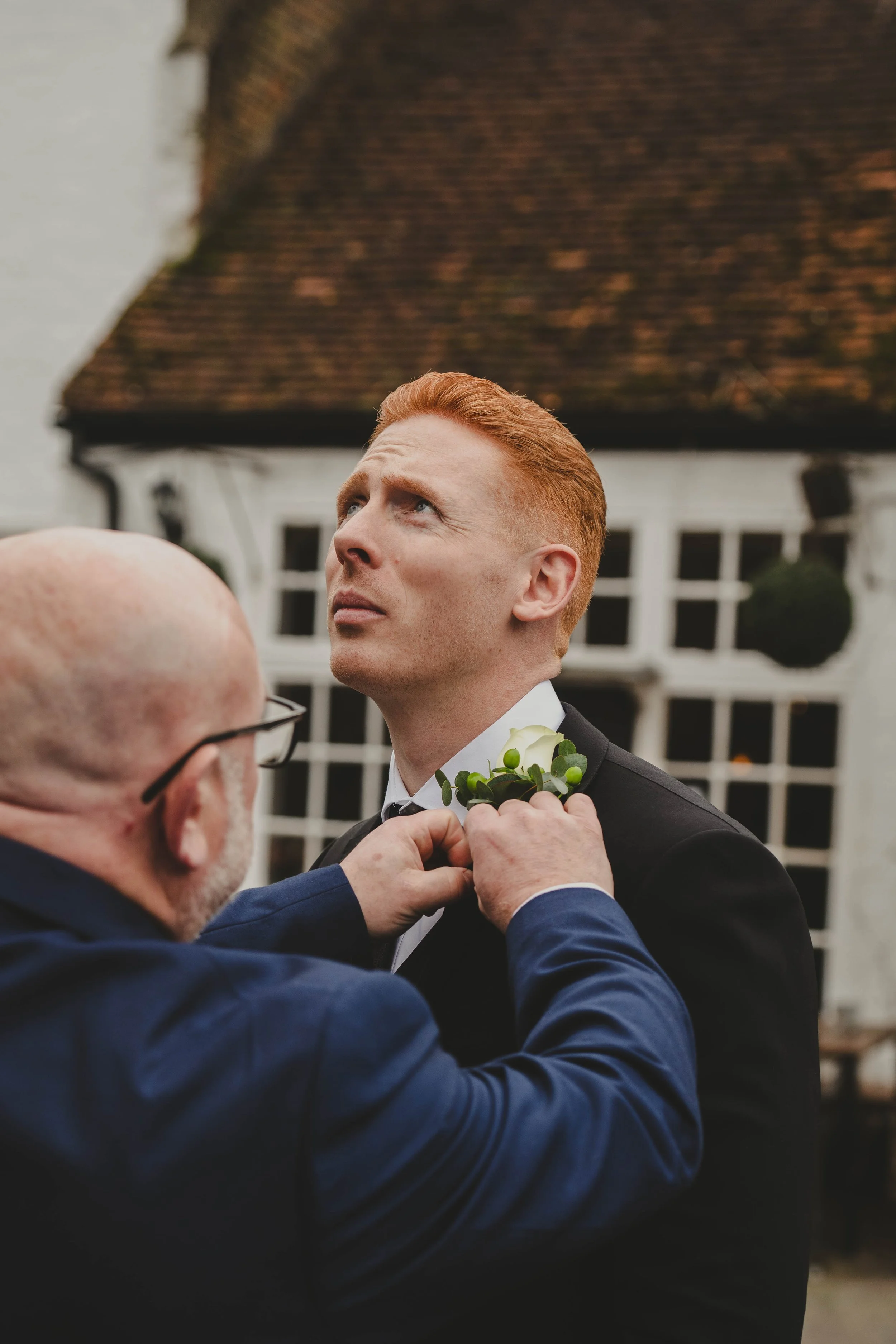 A man with red hair and a tuxedo is being helped with a boutonniere by another man in a blue suit at an outdoor wedding.