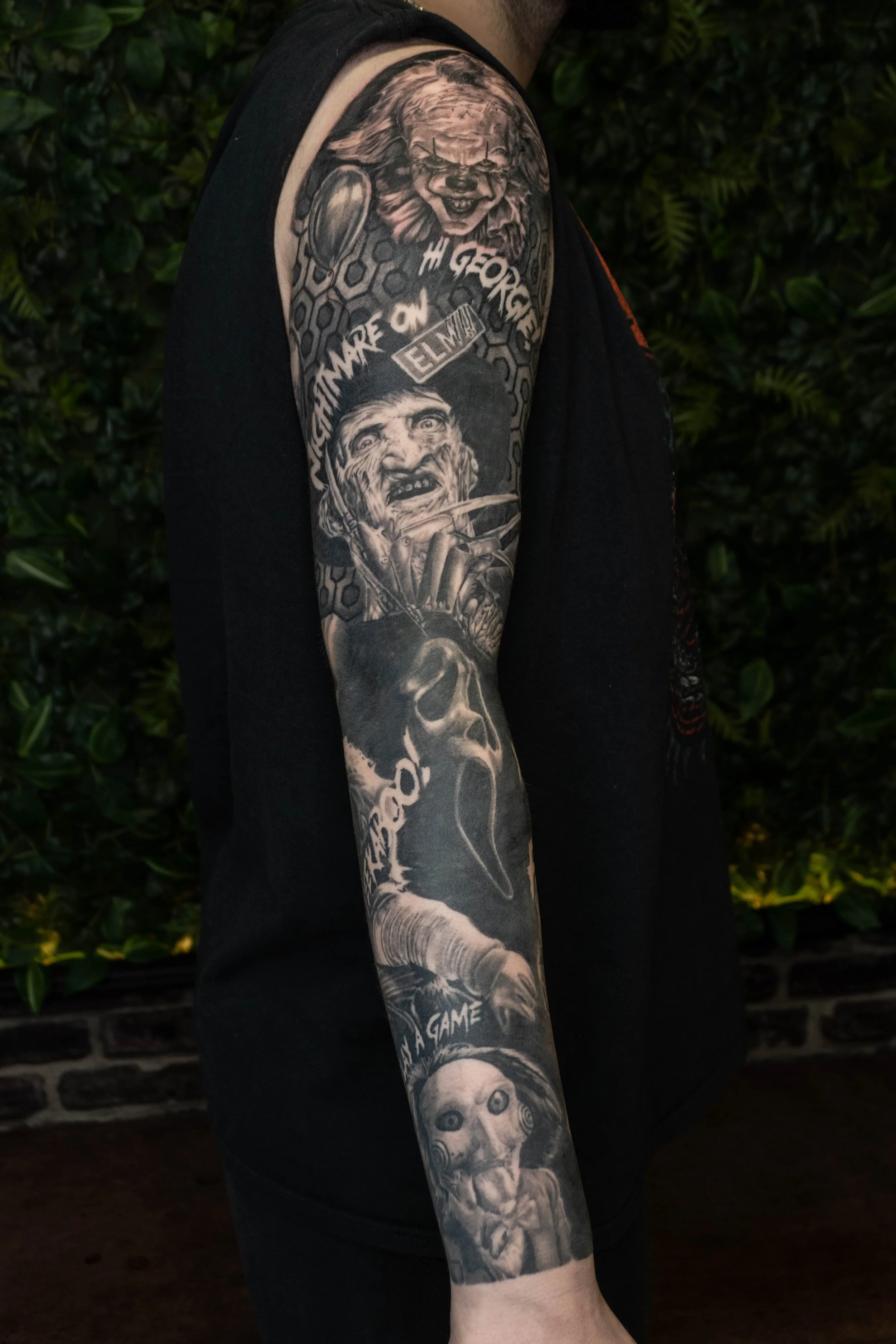 Black and grey sleeve tattoo featuring two characters: a clown or joker with an exaggerated face at the top and a detailed portrait of a woman with long hair holding a sword at the bottom, with various text and patterned background elements.