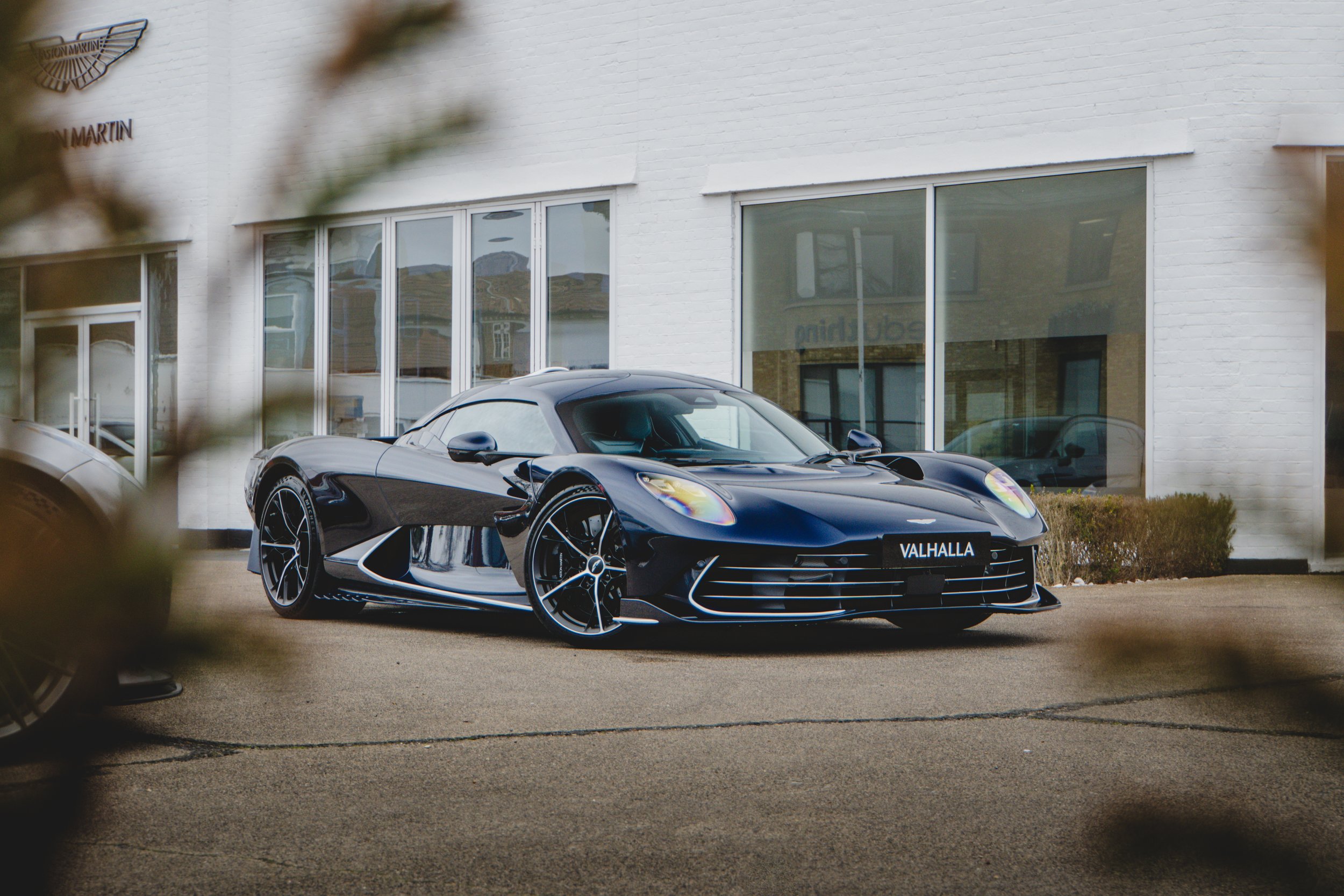 A black sports car labeled 'Valhalla' parked outside a commercial building with large windows. The building has a white brick exterior and a sign with an emblem and text partially visible.