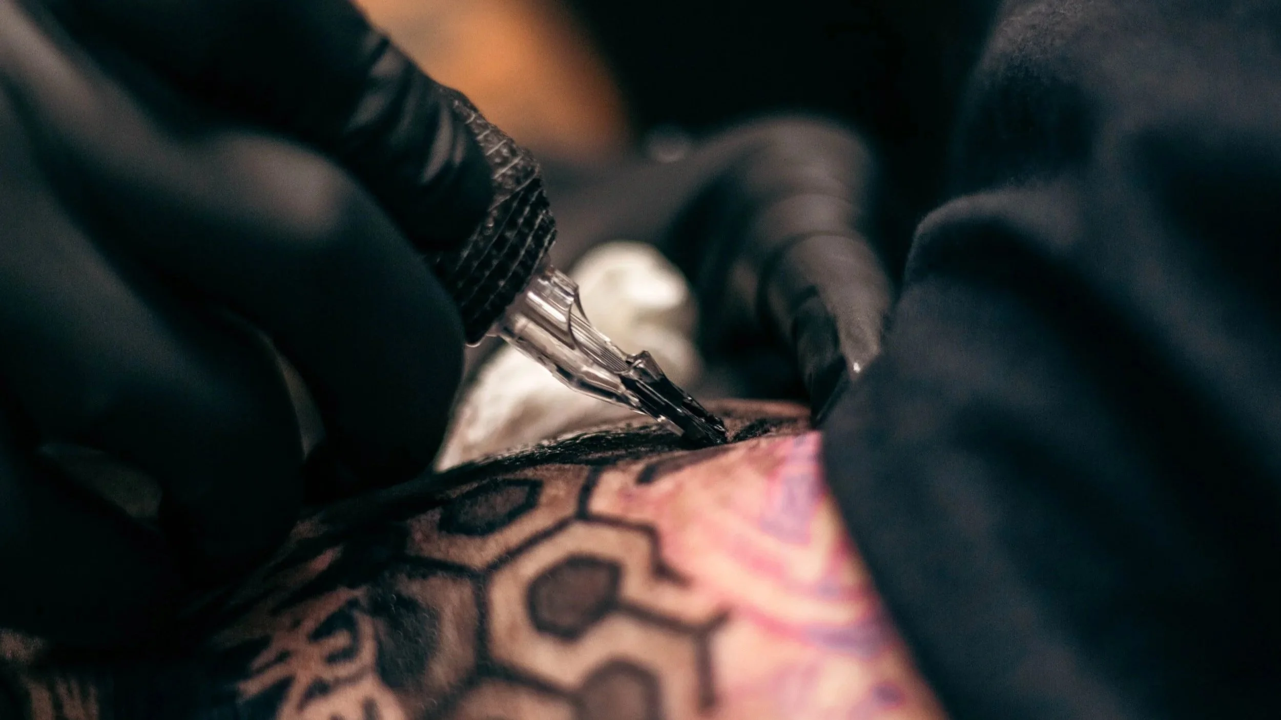 Close-up of a tattooed person's skin being tattooed with ink and needles by a tattoo artist wearing black gloves.