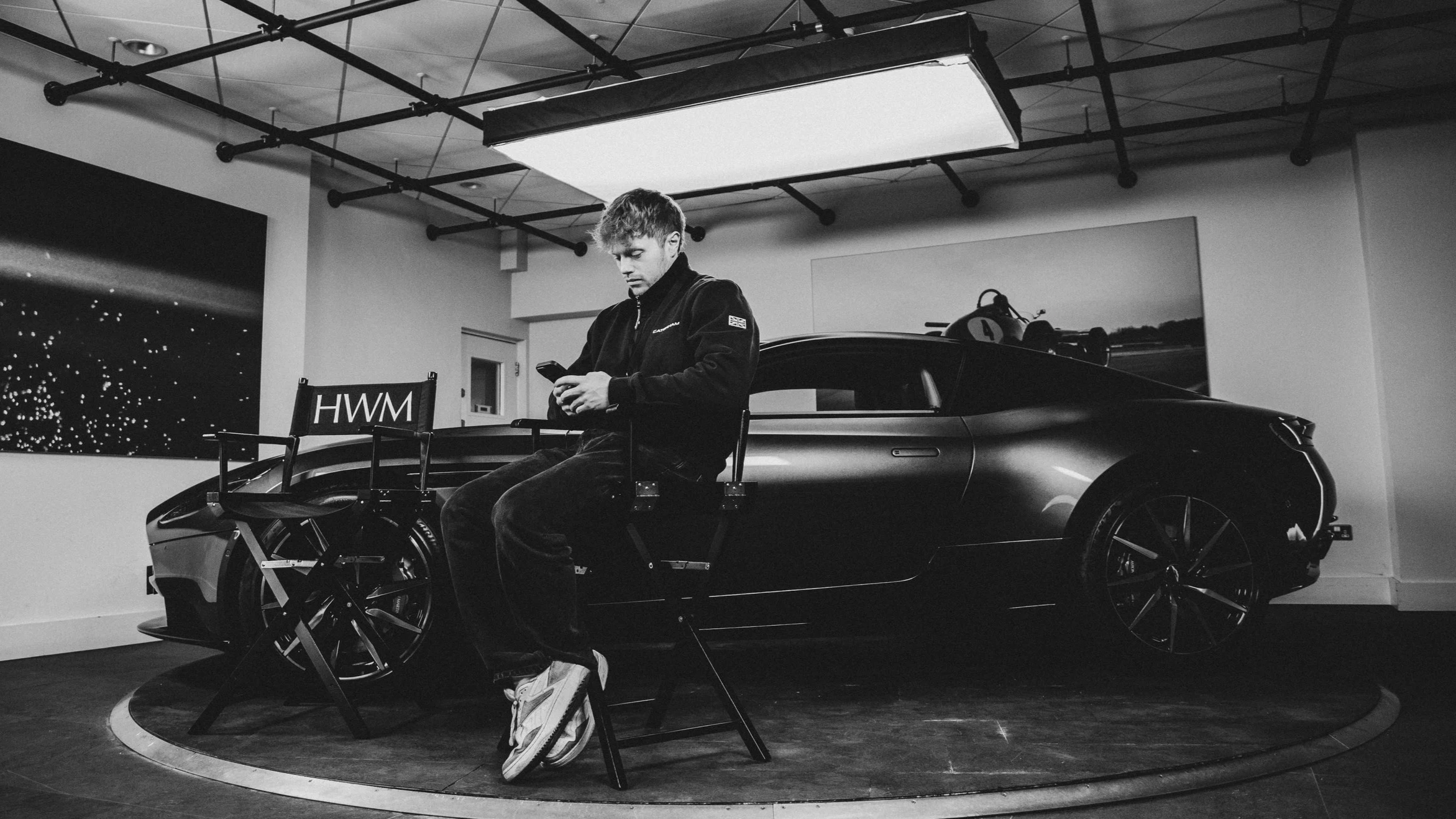 A man sitting on a chair and looking at his phone in a room with a sports car behind him, ceiling lights, and wall art.