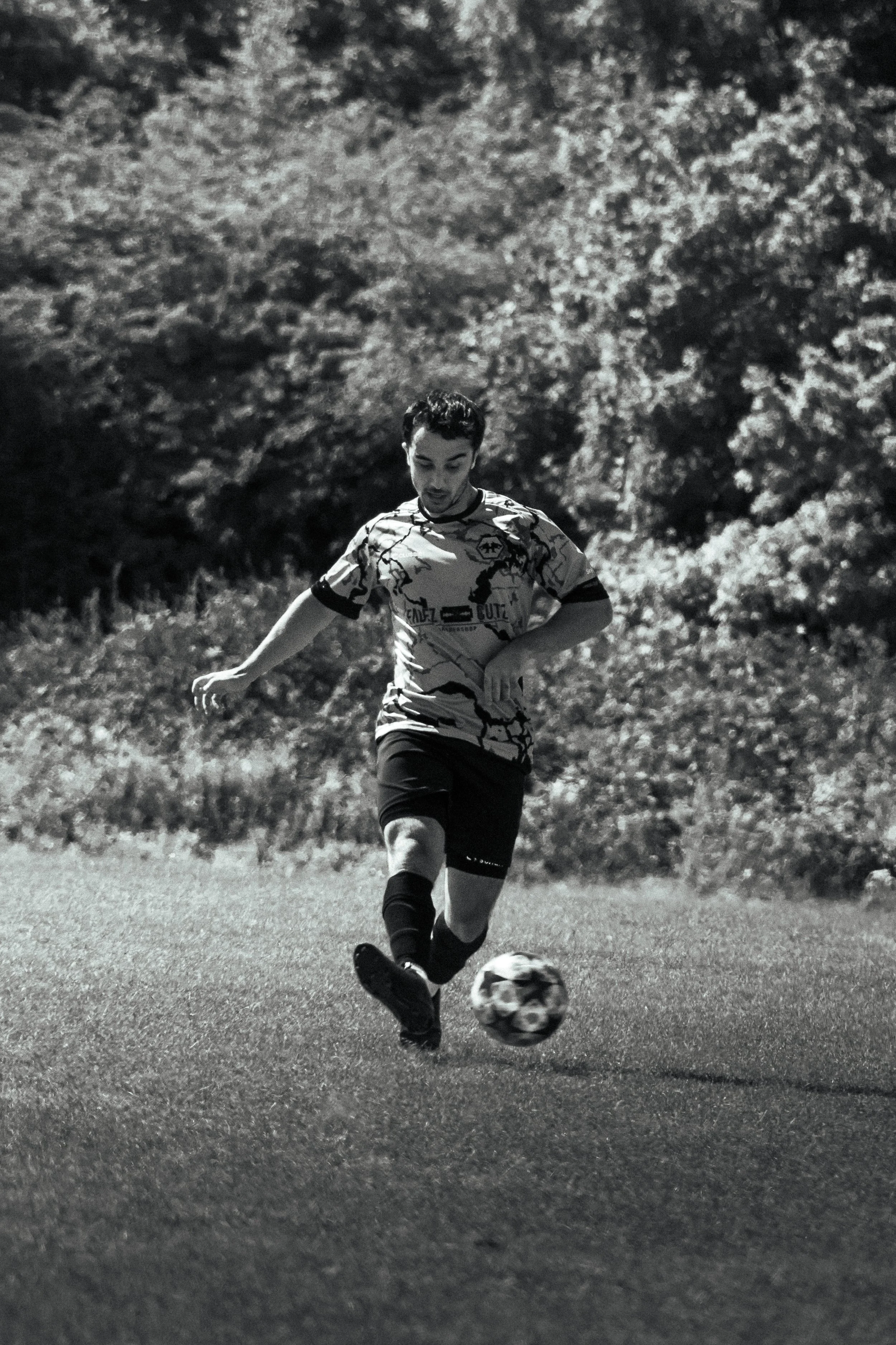A young man playing soccer outdoors on a grassy field, kicking a ball, with trees in the background.