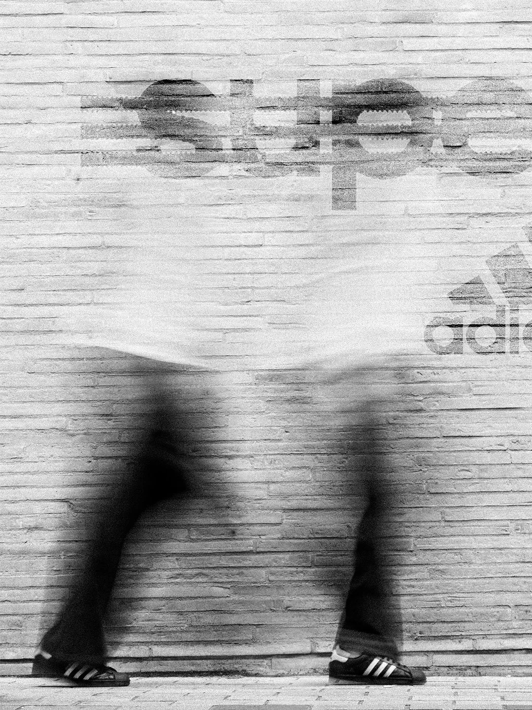 Person with blurred face in athletic clothing standing against a brick wall with a large Adidas logo.