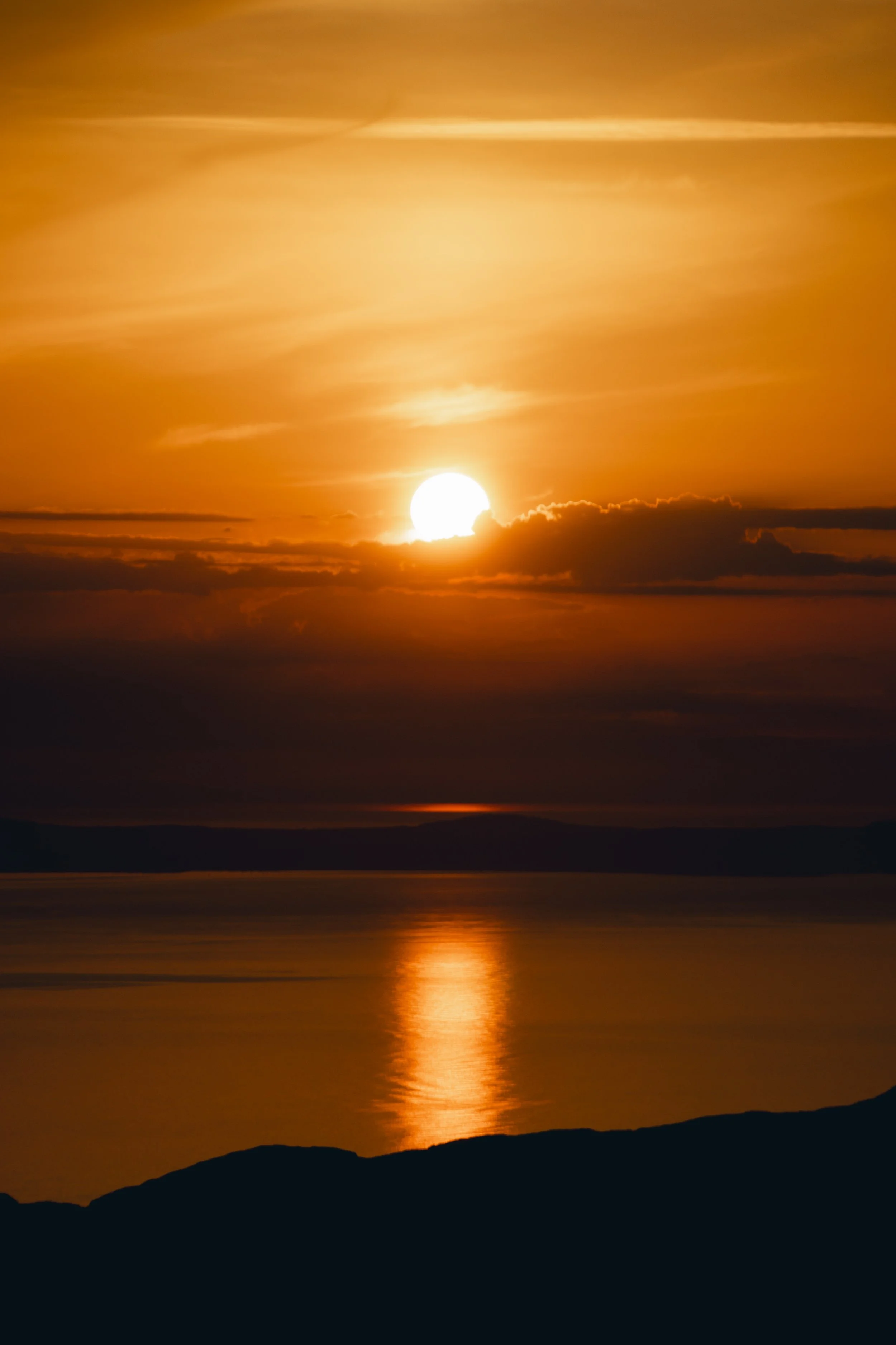 Sunset over a body of water with a reflection of the sun, clouds, and distant shoreline in a warm orange and yellow sky.