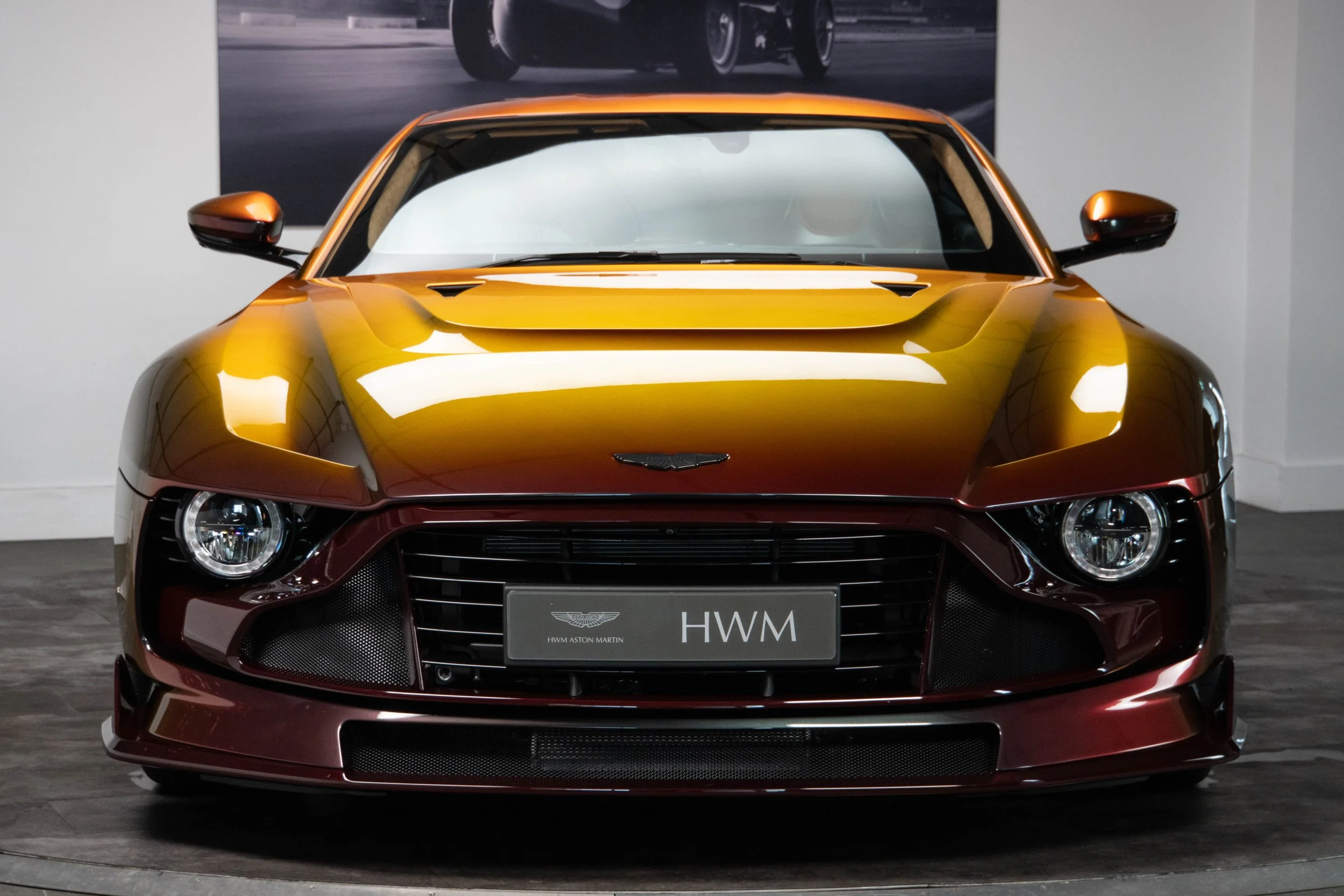 Front view of a yellow and burgundy Aston Martin car, displayed indoors with a large photograph of a car racing on the wall behind it.