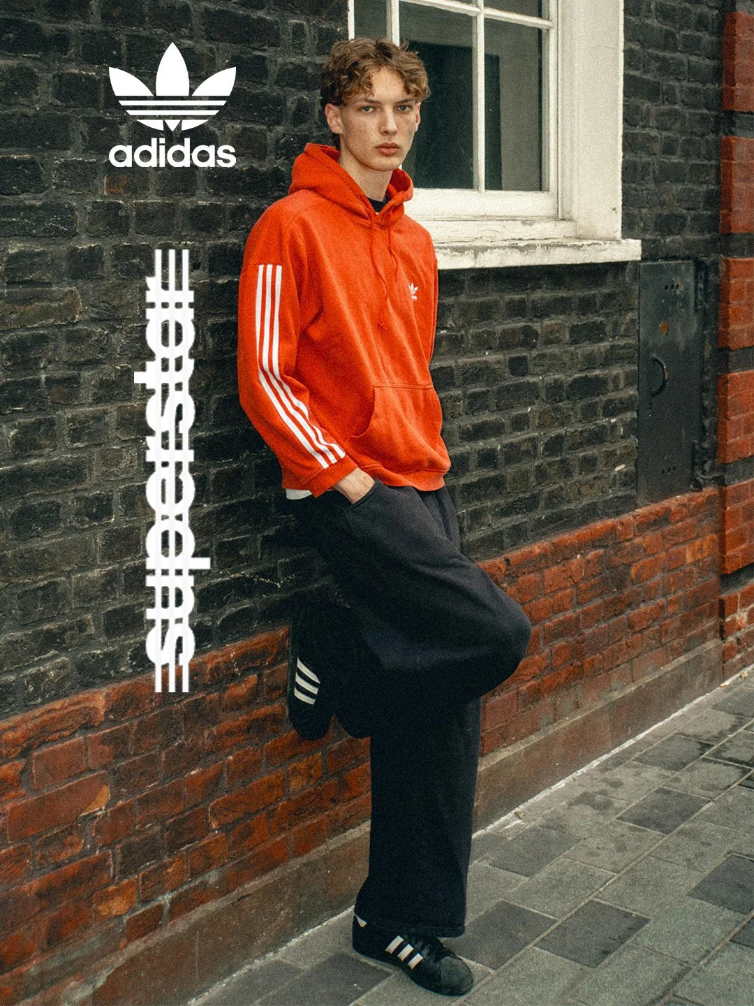 A young man leaning against a brick wall outside, wearing a bright red Adidas hoodie, black Adidas track pants, and black Adidas sneakers with white stripes, next to white Adidas logo and text.