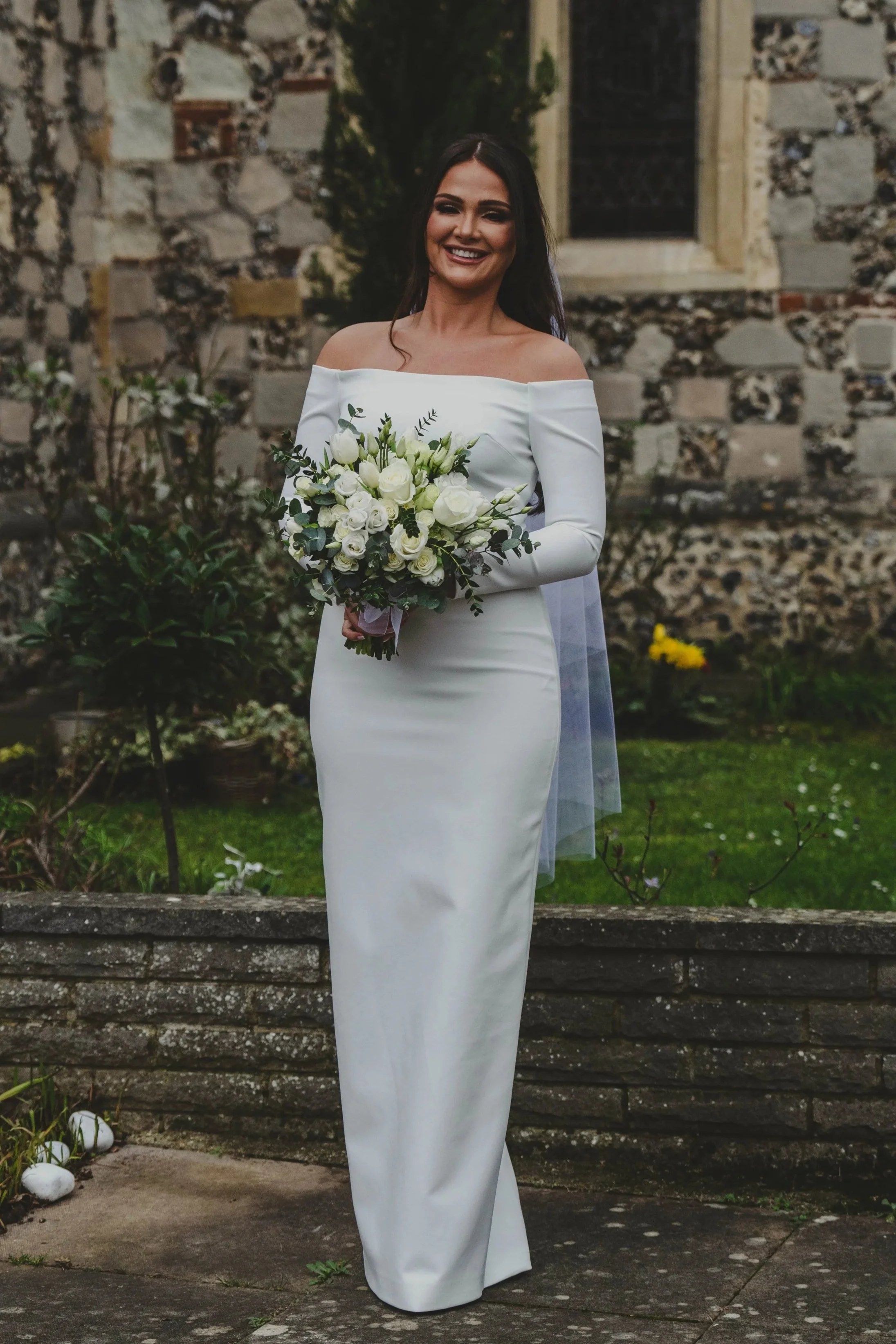 A woman in a white wedding dress holding a bouquet standing outdoors in front of an stone wall with plants.