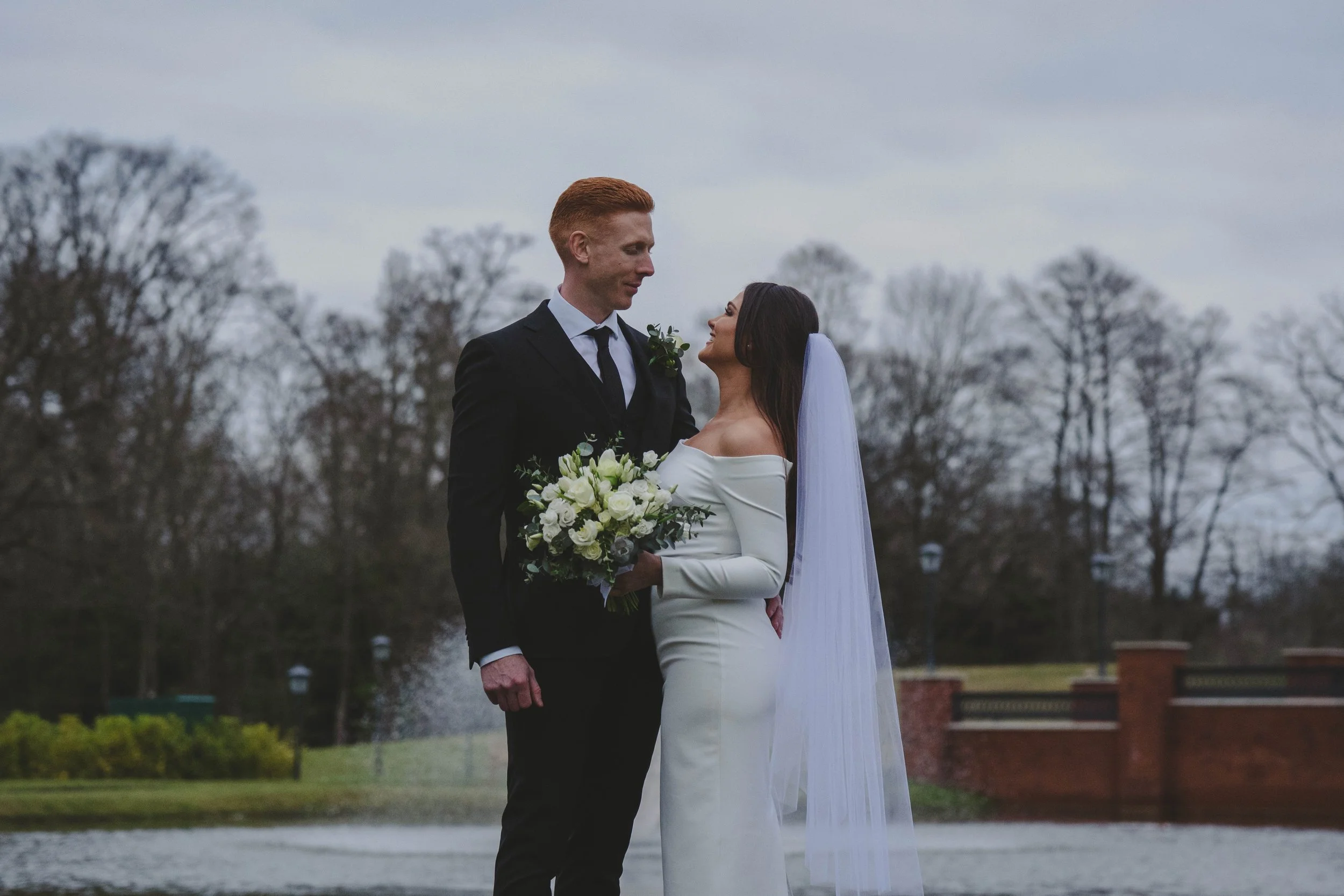 A bride and groom stand outdoors during a wedding ceremony, facing each other and holding hands, with trees and a park in the background on a cloudy day.
