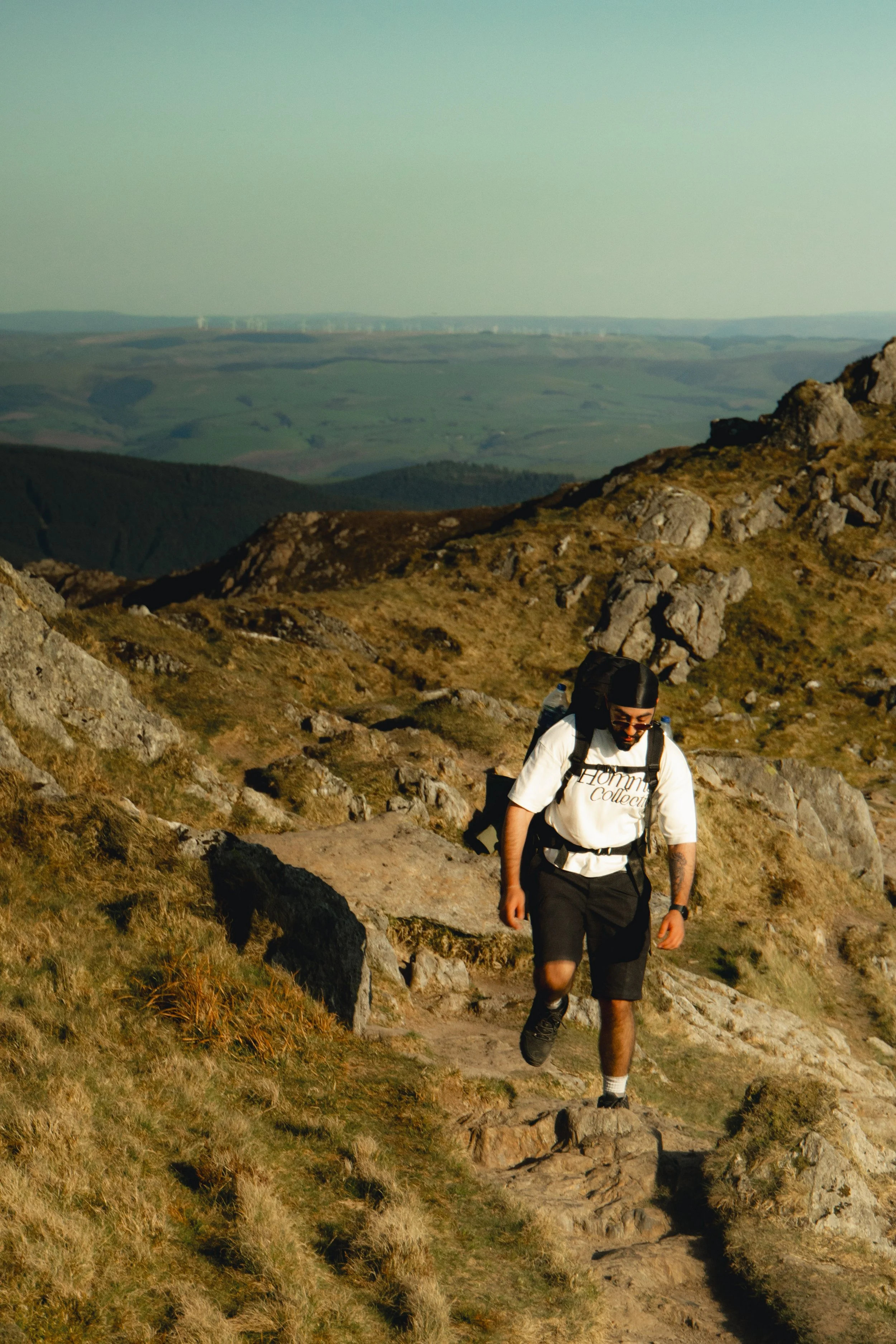 A person hiking up a rocky trail on a mountain during sunset, with rolling hills and wind turbines in the distance.