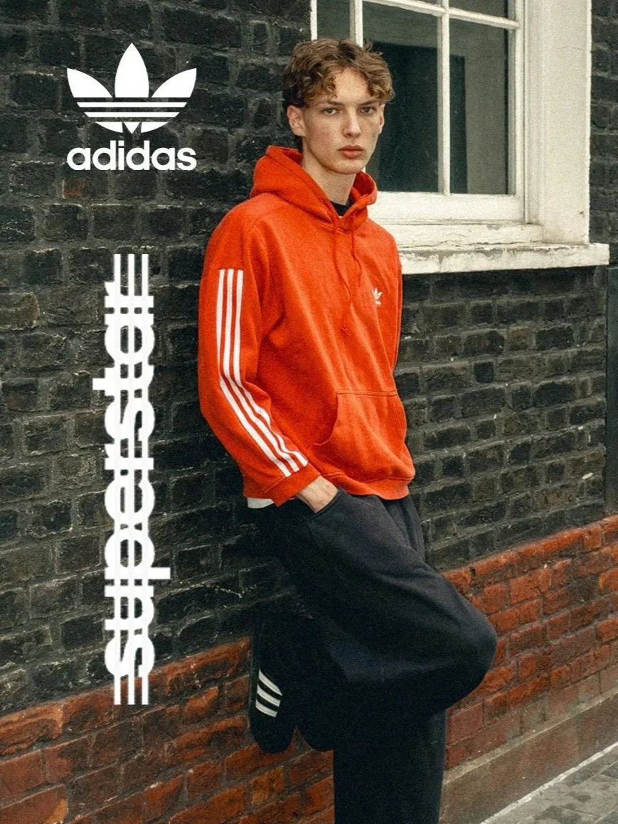 A young man wearing a red Adidas hoodie with white stripes and black pants, leaning against a brick wall near a window.