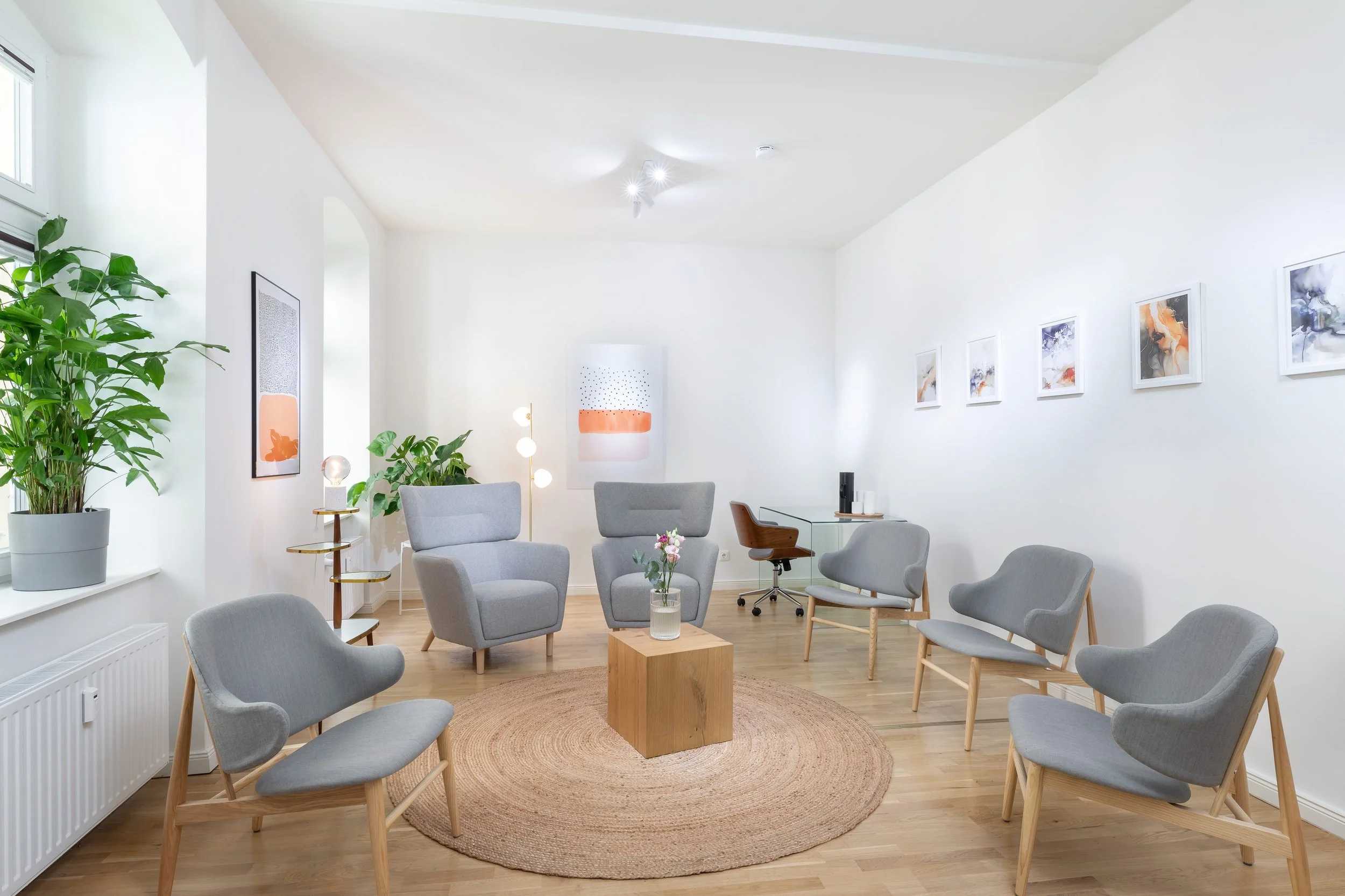 Helle, moderne Wohnzimmer mit mehreren grauen Stühlen, einem kleinen Holztisch mit Vase und Blumen, Kunstwerken an den weißen Wänden, Zimmerpflanzen, Holzfußboden und einer runden Teppich im Zentrum.