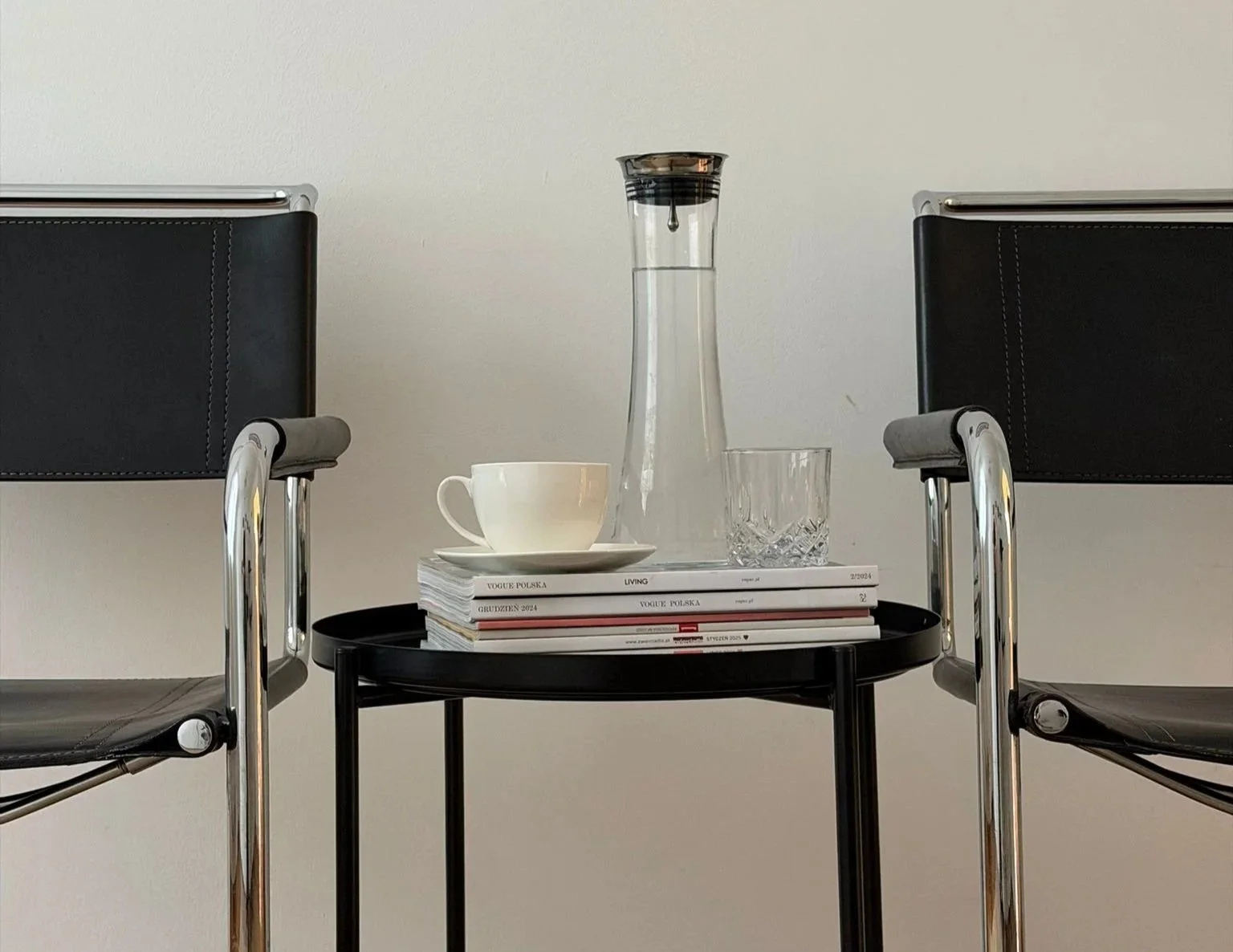Two black chairs with chrome frames facing each other, a small black table between them with magazines, a white tea cup and saucer, a clear glass water pitcher, and a drinking glass on top, against a plain light-colored wall.