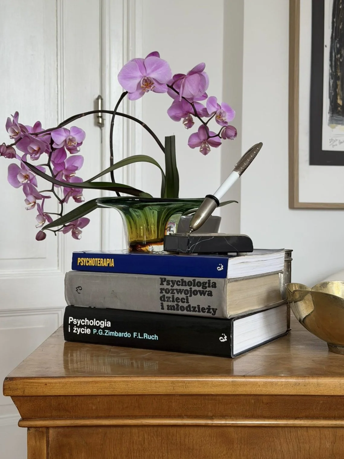 A stack of four books on a wooden table with an orchid plant, a pen, and a brass bowl on top. The books are titled 'Psychoterapia', 'Psychologia rozwojowa dzieci i młodzieży', and 'Psychologia i życie'.