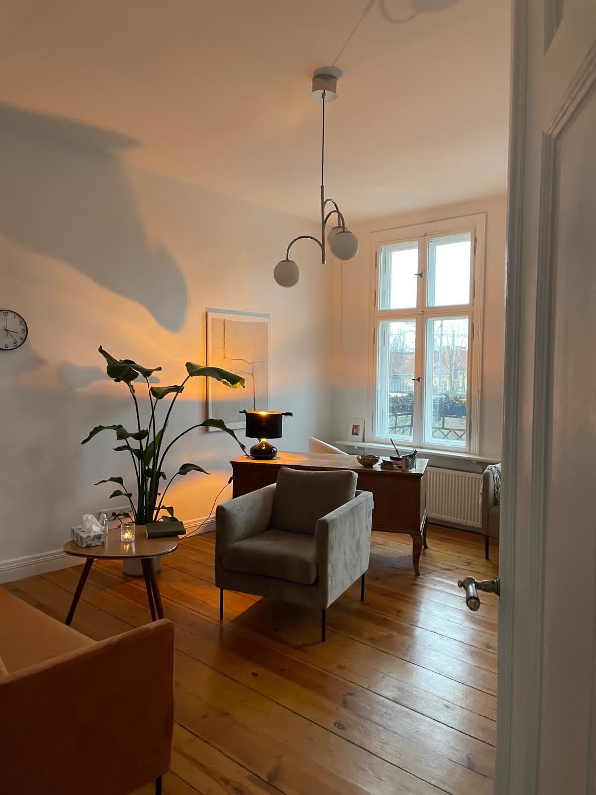 A cozy living room with a large window, wooden floors, a beige armchair, and a wooden desk decorated with a lit lamp, a bowl, and small items. A tall plant is near the wall, and a ceiling light hangs from the ceiling.