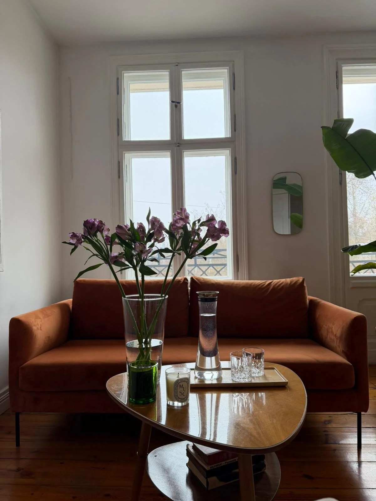 A living room with a wooden coffee table in front of a brown sofa, decorated with a glass vase of pink flowers, a tall glass carafe, candles, and glasses. Large windows with white frames are behind, letting in natural light. A small mirror and green 