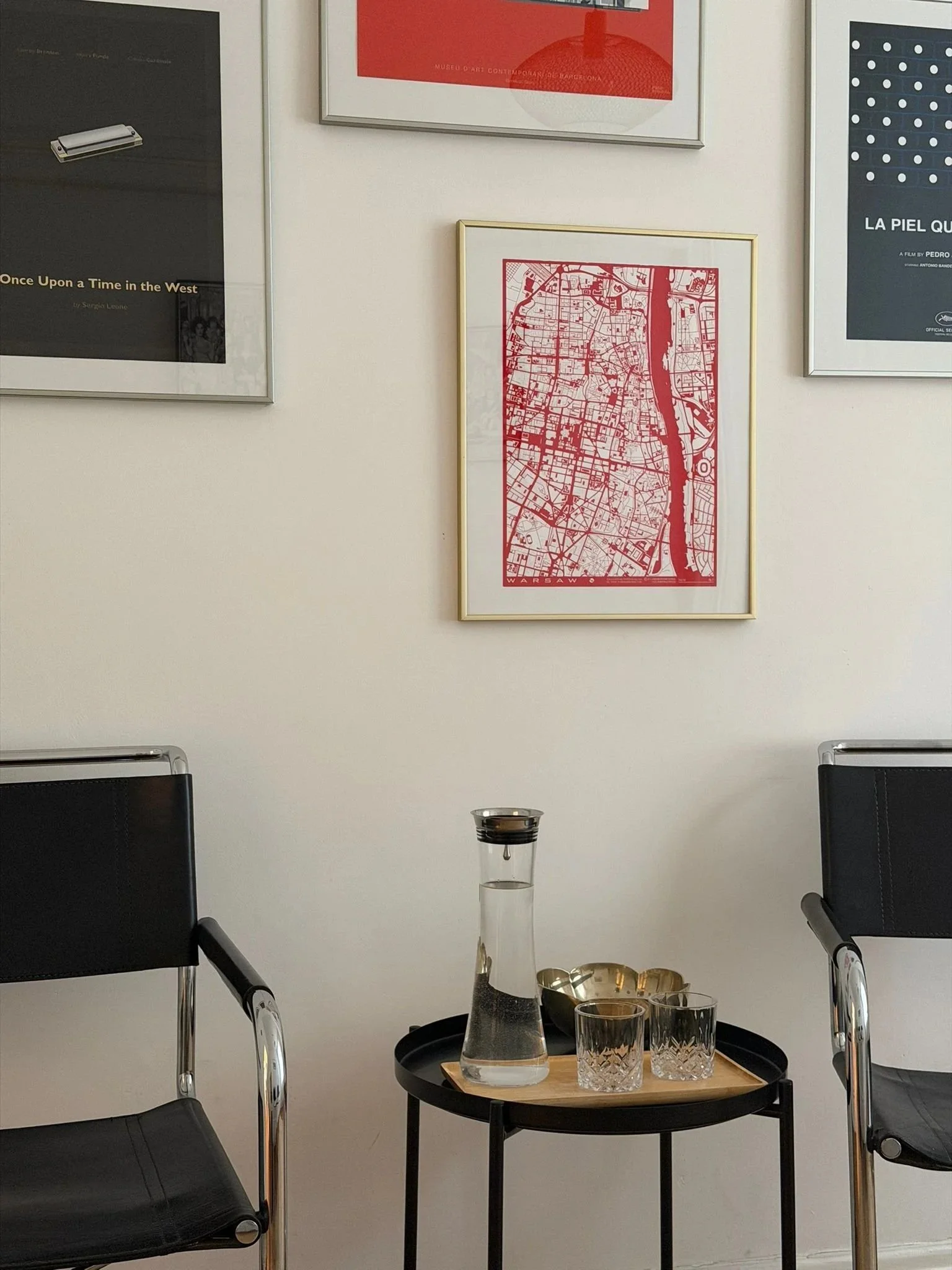 A minimalist dining area with a small black round table set with a glass pitcher, three empty glasses, and a wooden tray. Two black chairs with chrome legs are positioned on opposite sides of the table. The background features a white wall with three