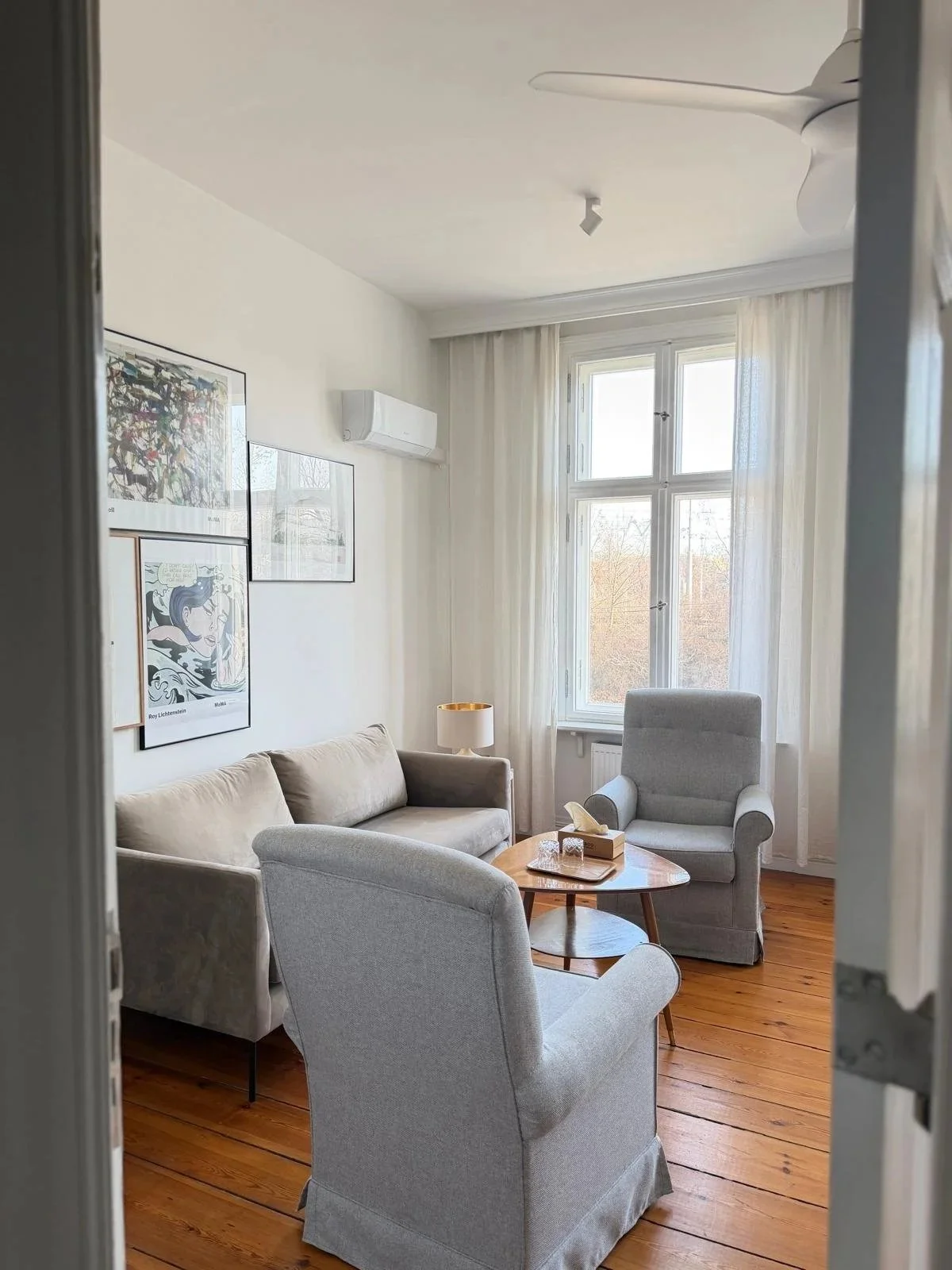 A cozy living room with three beige armchairs, a light-colored sofa, a wooden coffee table with a tissue box and glasses, a small table lamp, large window with white curtains, artwork on the wall, and wooden floor.