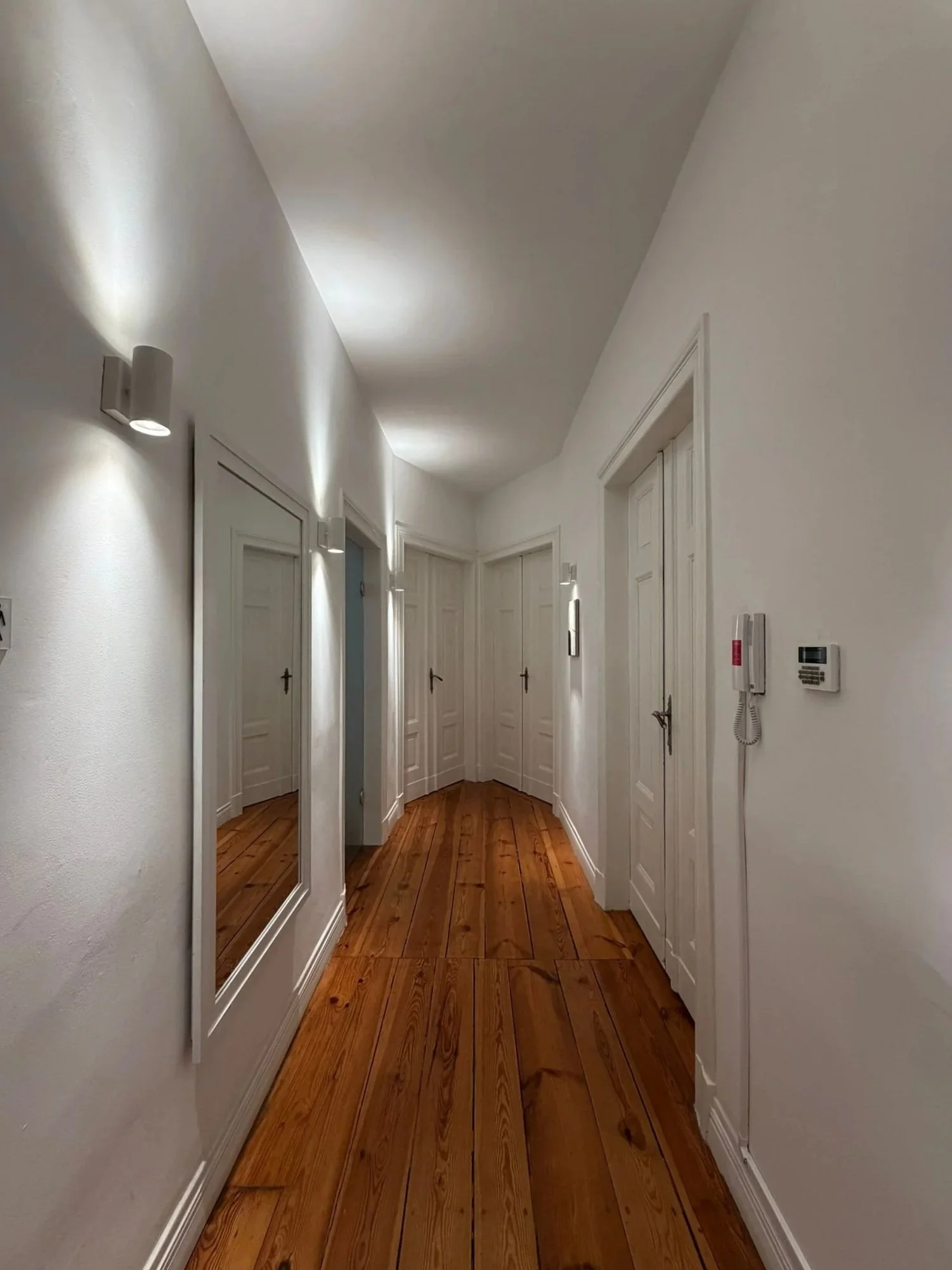 Hallway with white walls, wooden floor, and multiple closed white doors. Wall-mounted lights illuminate the hallway, and a large mirror is on the left side. On the right wall, there is a wall-mounted phone and control panel.