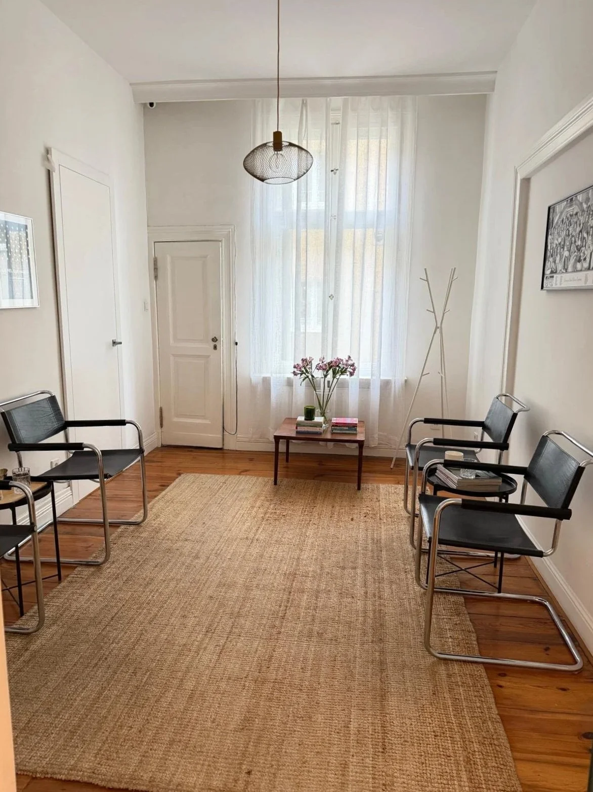A waiting room with four black chairs with metal armrests arranged two on each side, a wooden coffee table with flowers and books, a beige rug, white walls, a large window with sheer curtains, a ceiling light, a coat rack, and framed artwork on the w