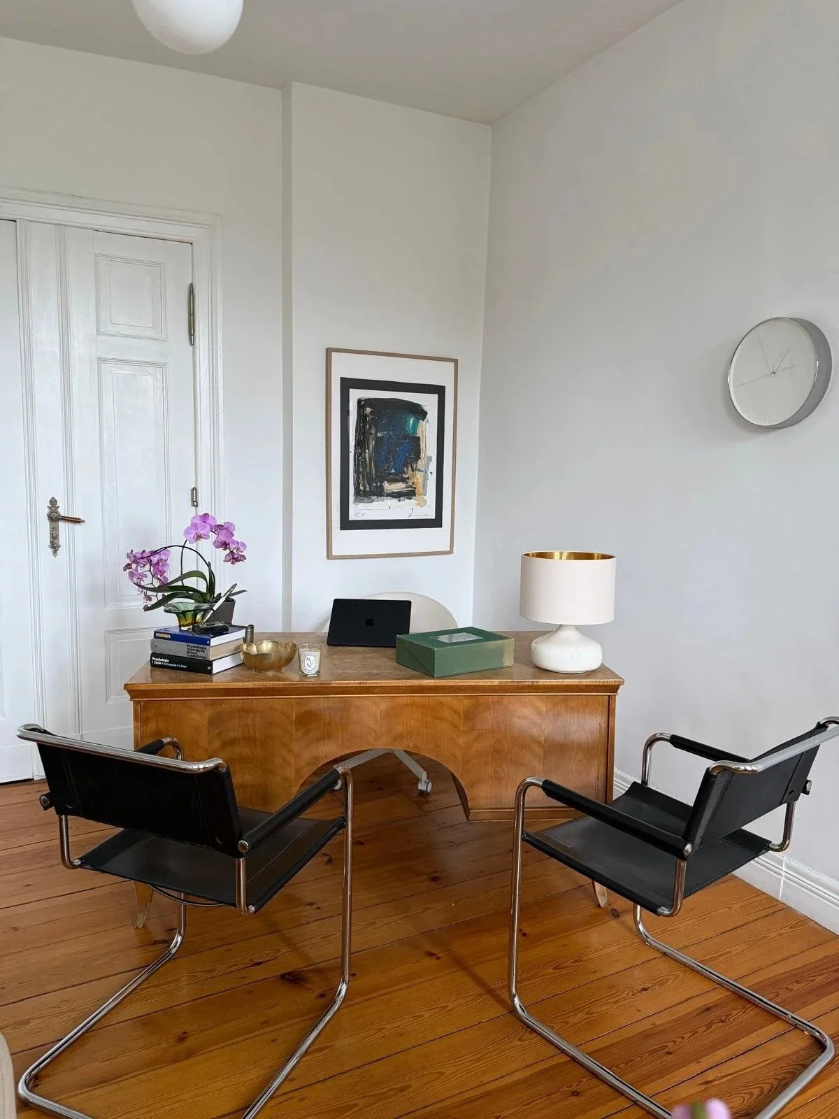 Office space with a wooden desk, two black chairs, a white lamp, a potted orchid, books, and artwork on a white wall.