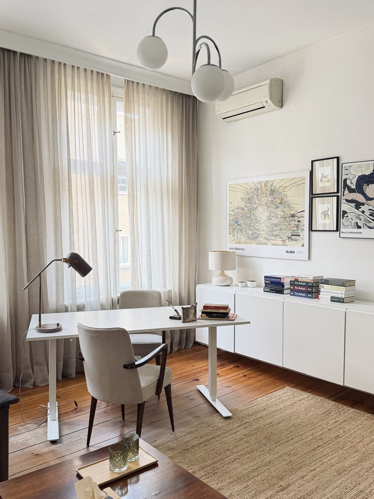 Bright, modern home office with white desk, beige armchair, bookshelf with stacked books, framed artwork on wall, beige curtains, wooden floor, and ceiling light fixture.