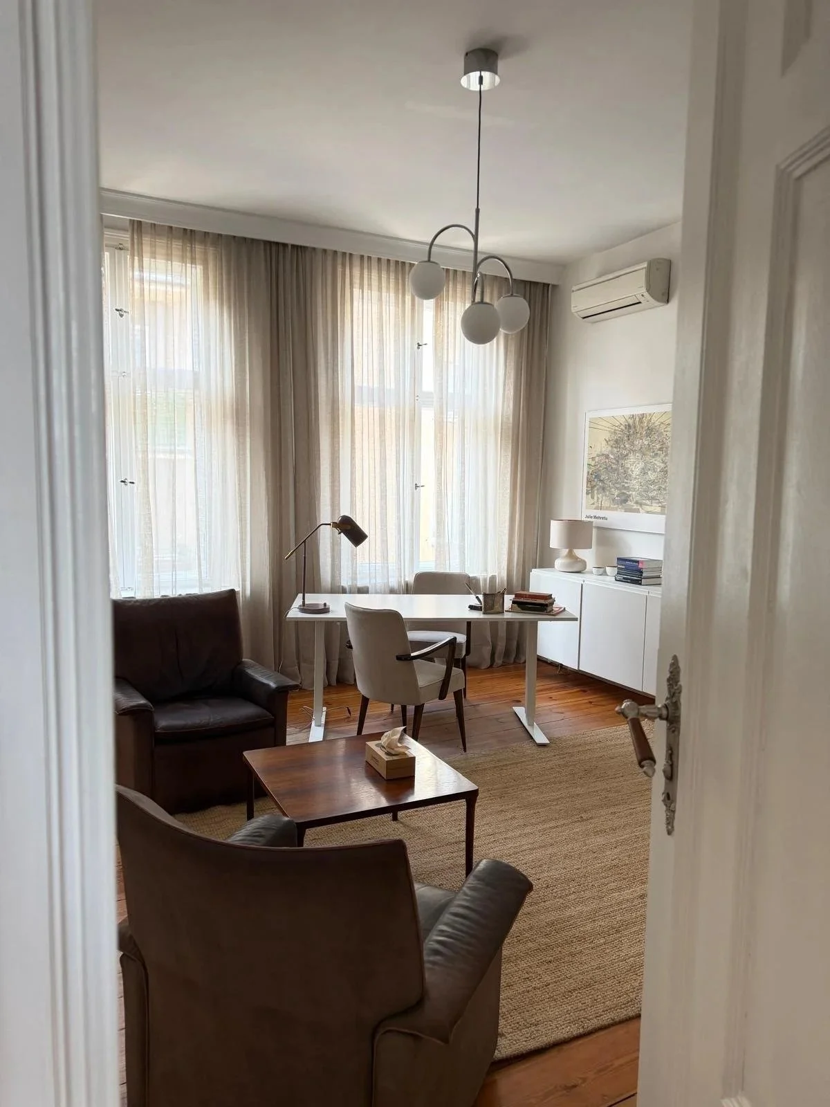 A well-lit home office with a white desk, beige armchair, brown leather sofa, and two white cabinets. The room has large windows with sheer beige curtains, a modern chandelier, a desk lamp, and a framed artwork on the wall.