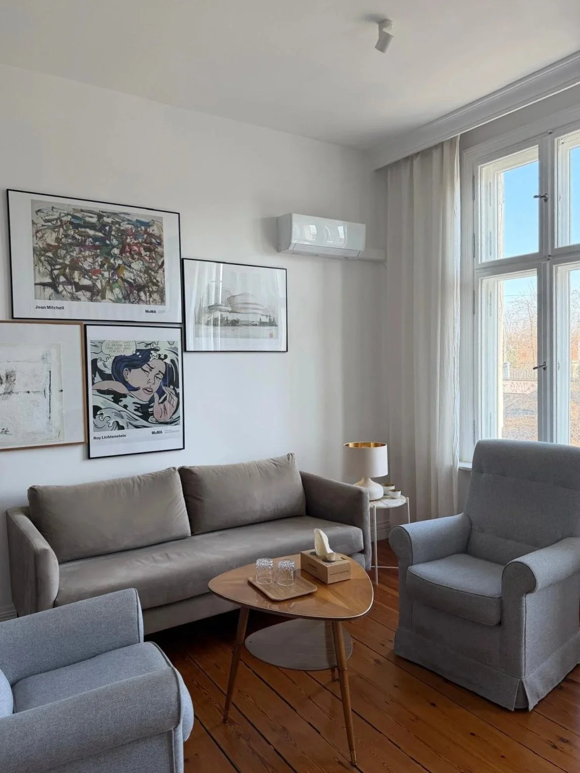 Living room with beige sofa, two armchairs, wooden coffee table, white side table with lamp, wall art, and large windows with curtains.