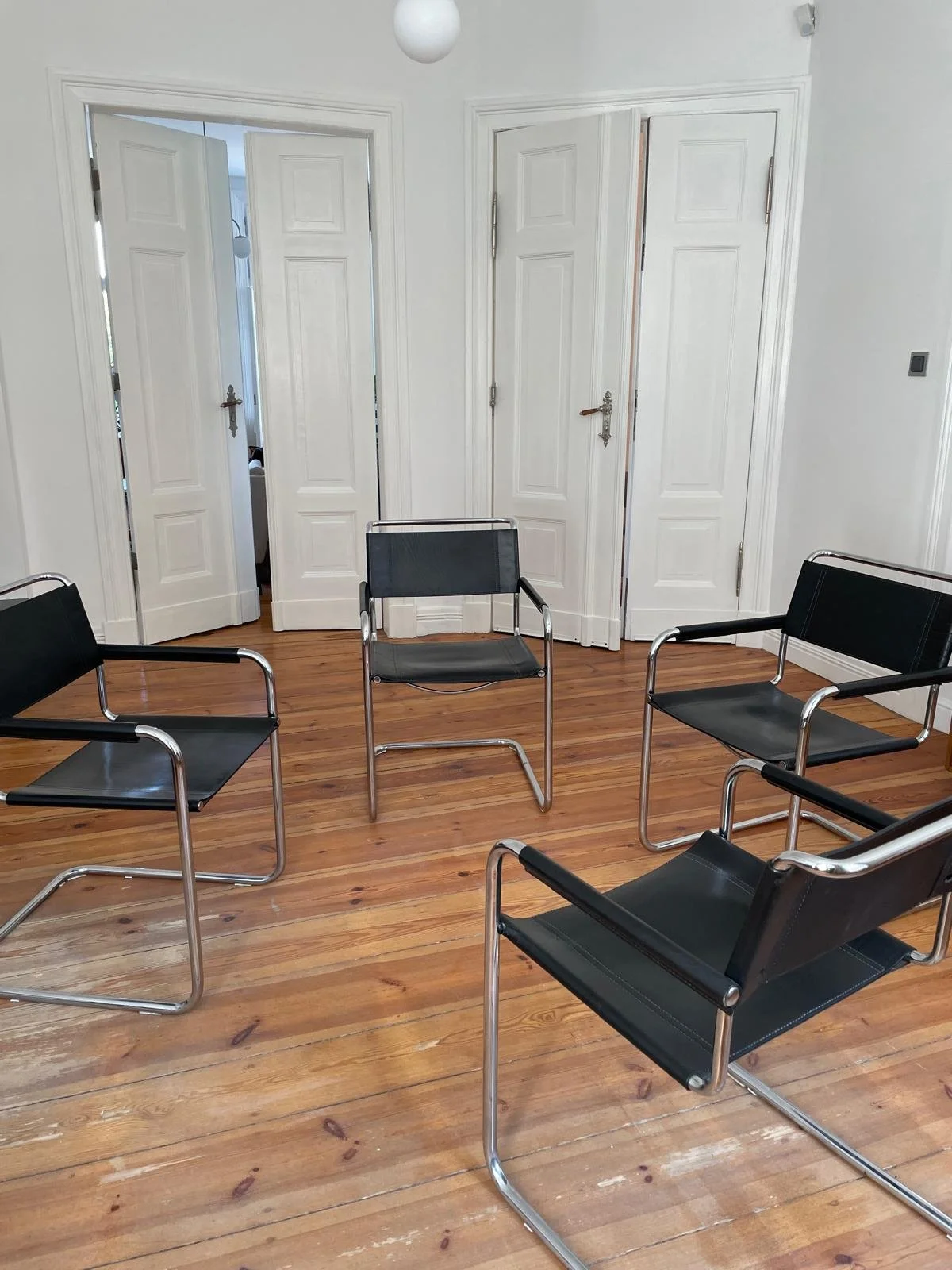 A room with wooden floors and white walls, containing five black and metal chairs arranged in a circle. There are two closed white closet doors, one open, with a visible adjacent room. A white ceiling light is hanging, and a light switch is visible o