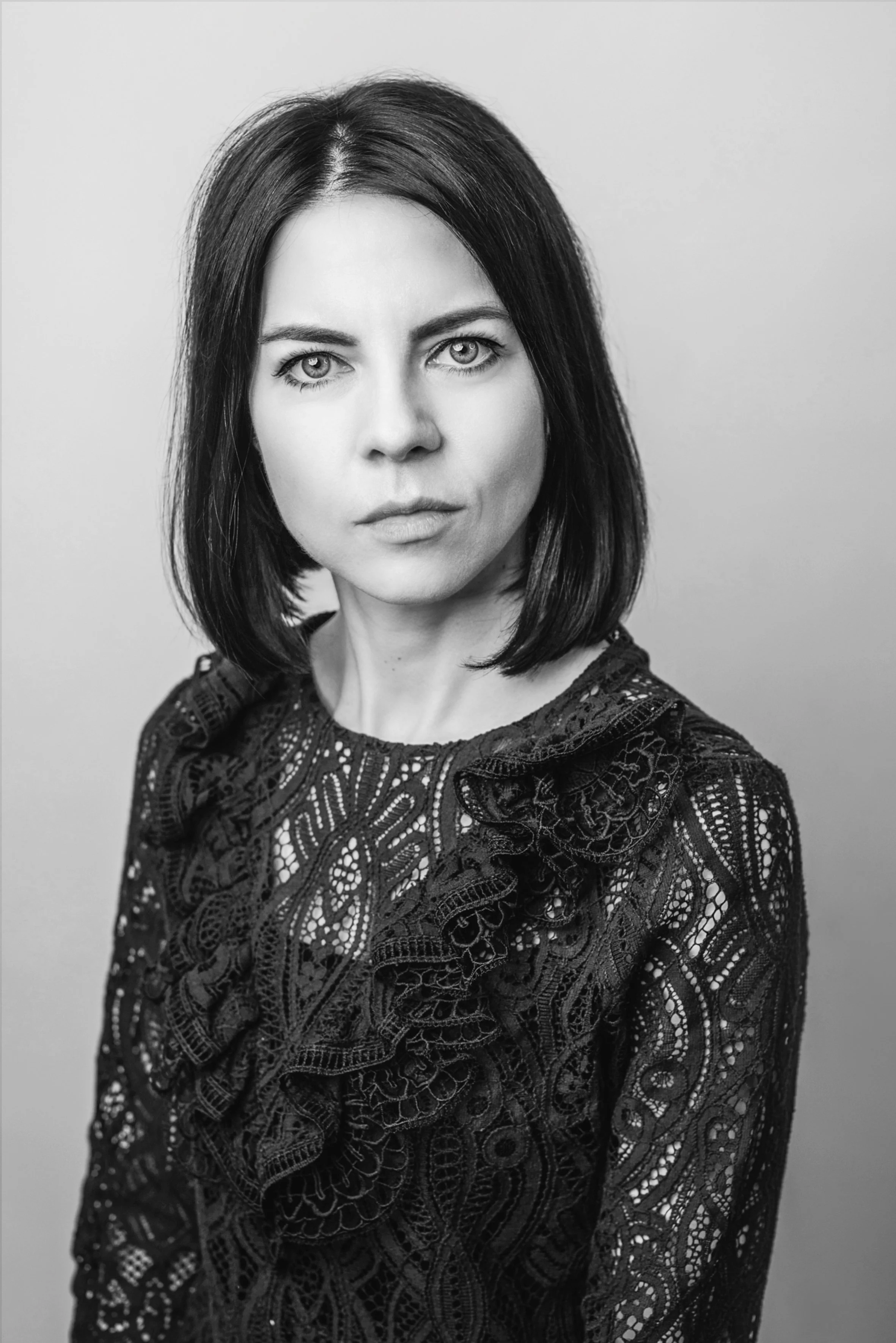 Black and white portrait of a woman with straight, shoulder-length dark hair, wearing a lace top with intricate patterns.
