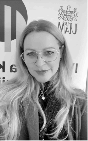 Black and white photo of a woman with long, wavy hair wearing glasses and a dark top, standing in front of a wall with logos and designs.