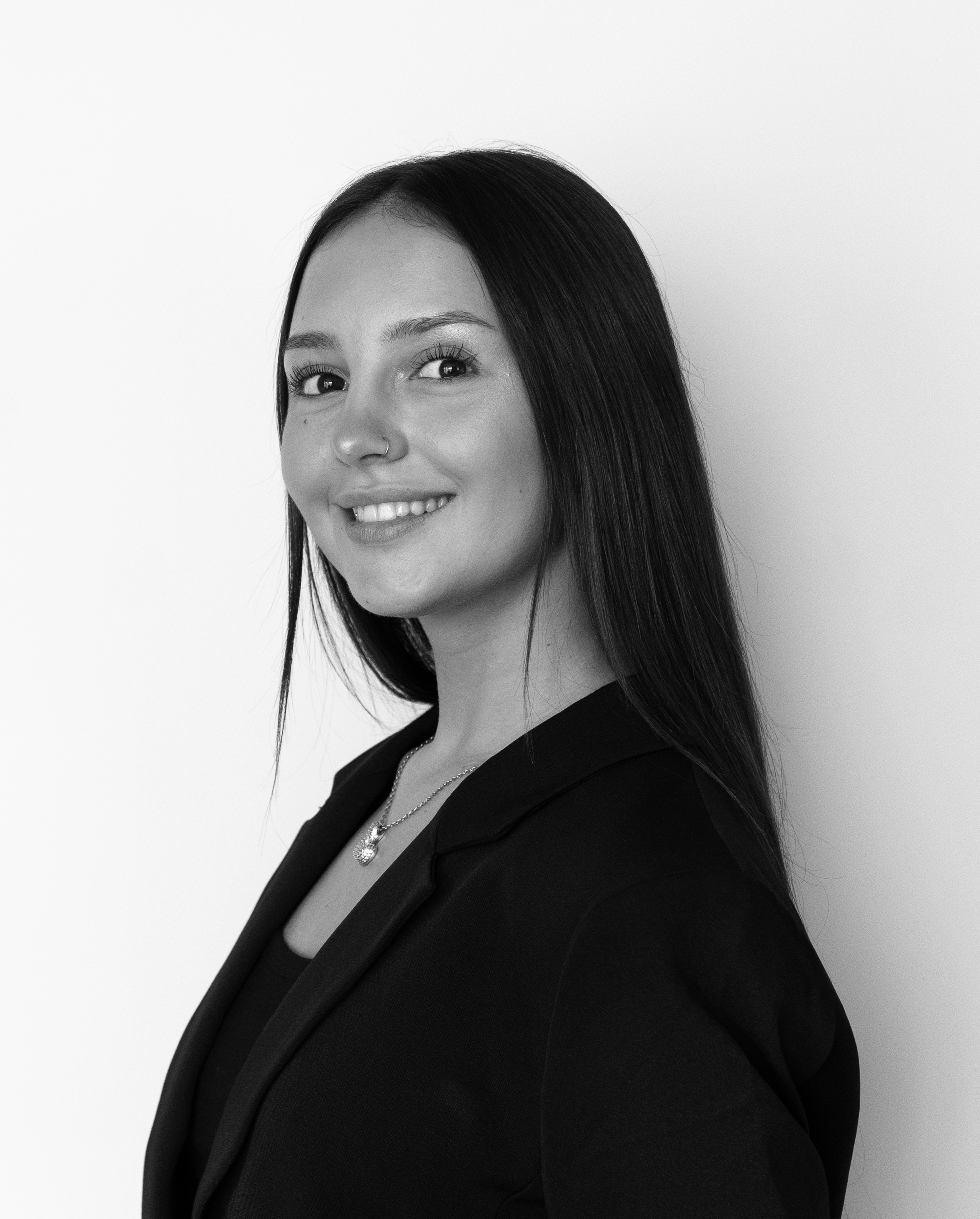 Close-up black and white portrait of a smiling woman with long dark hair, wearing a black blazer and a necklace, against a plain light background.