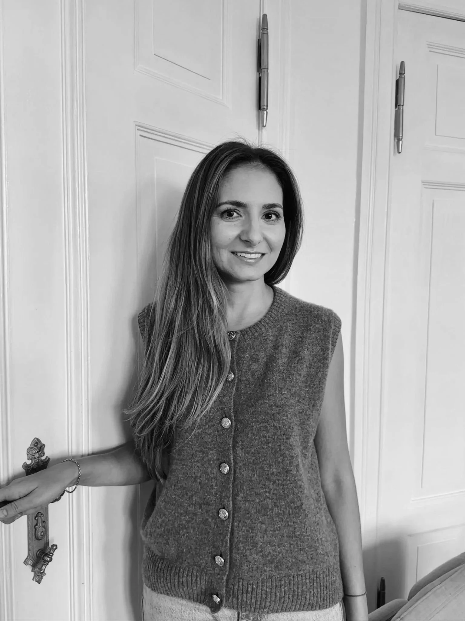A woman with long hair smiling, wearing a buttoned cardigan, standing indoors with a closet door in the background.