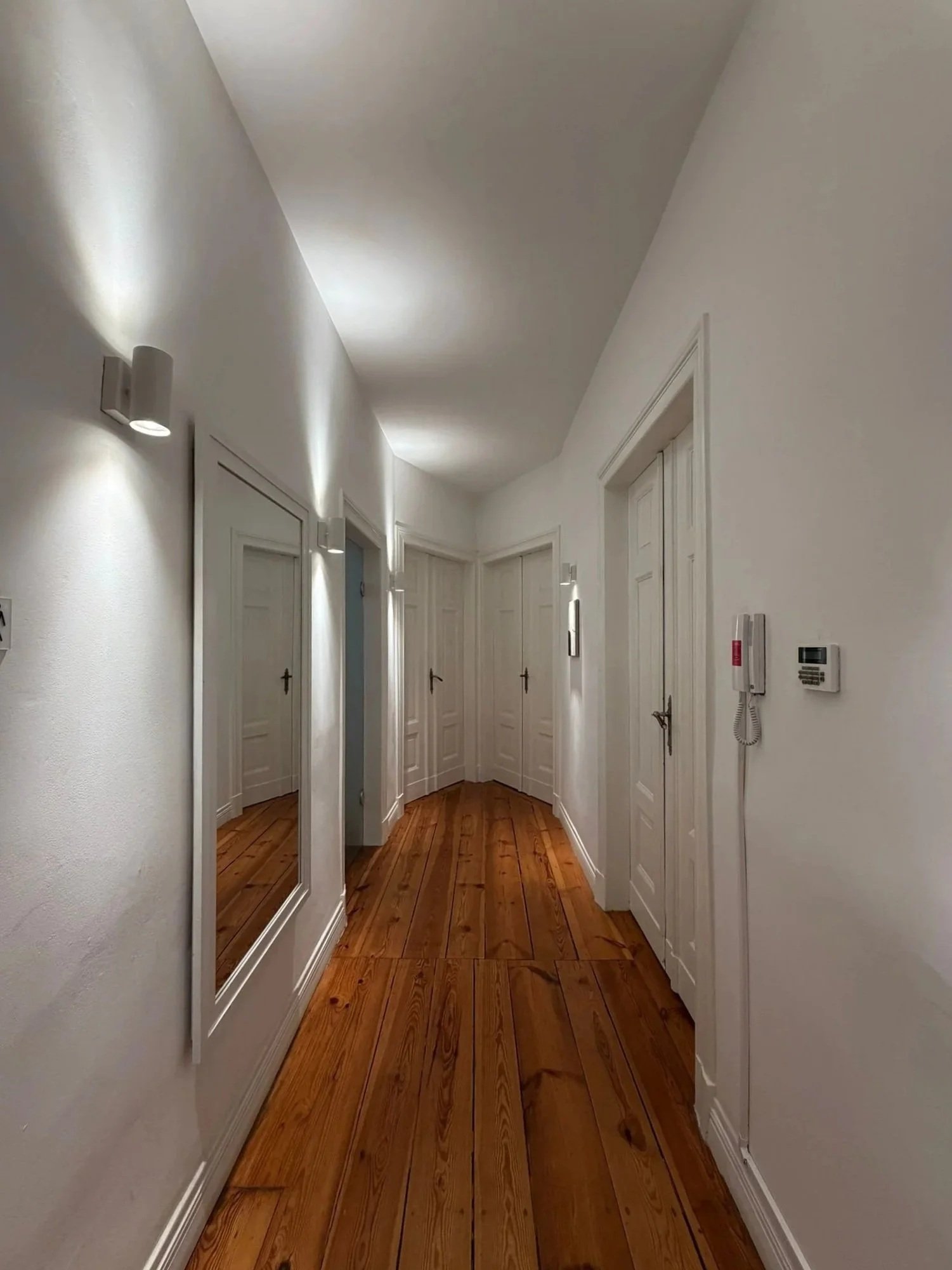 A hallway with white walls, wooden floors, several doors, a large mirror, wall-mounted lights, and a fire alarm system.