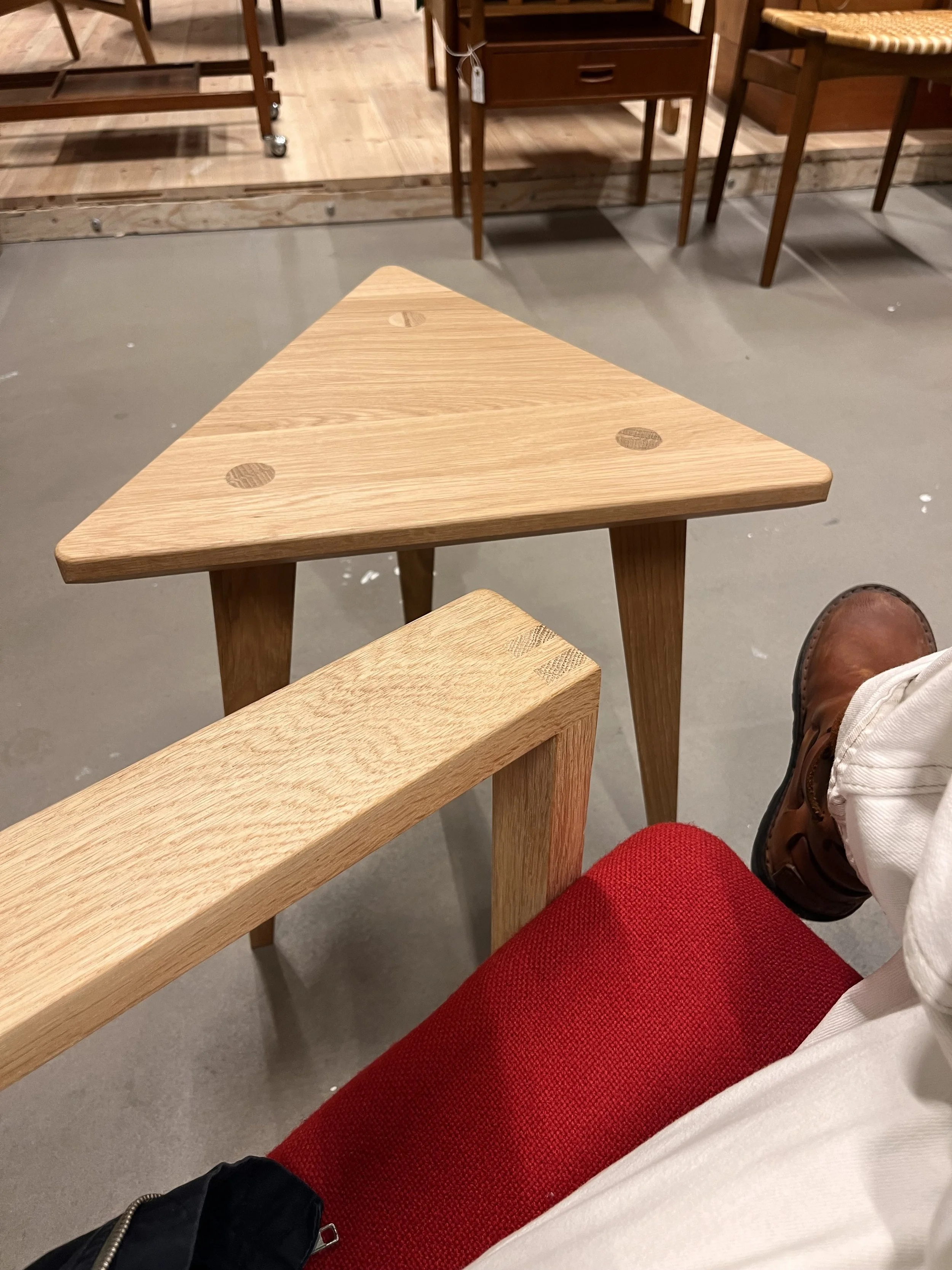 En person i vita byxor och röda byxor sitter på en stol i ett möbelbutik eller ett liknande utrymme, med ett rutnät av trästolar och möbler i bakgrunden.