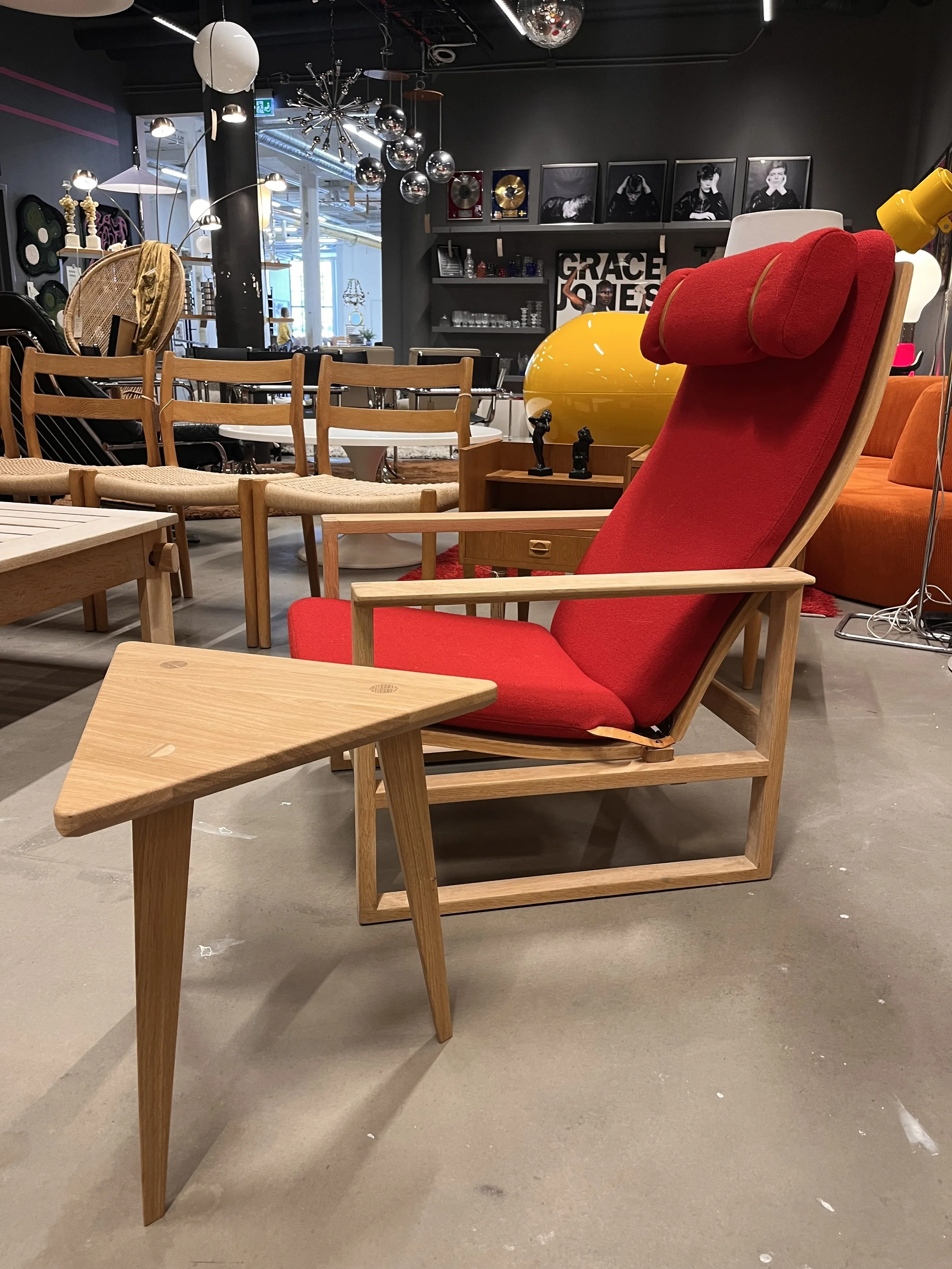En röd fotölj med träram, framför ett litet träbord, i ett inomhusrum med olika möbler och dekorationer, inklusive väggkonst, lampor och bilder.
