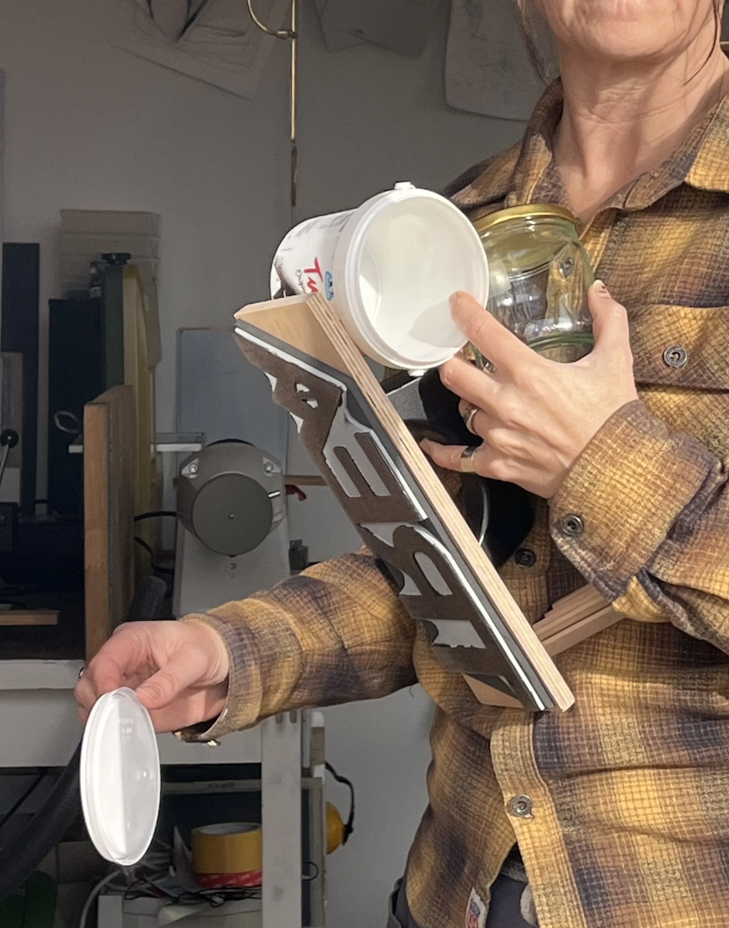 En person i rutnätsmönstrad skjorta som håller ett vitt plastlock till en burk eller behållare i ett verkstad eller arbetsrum.